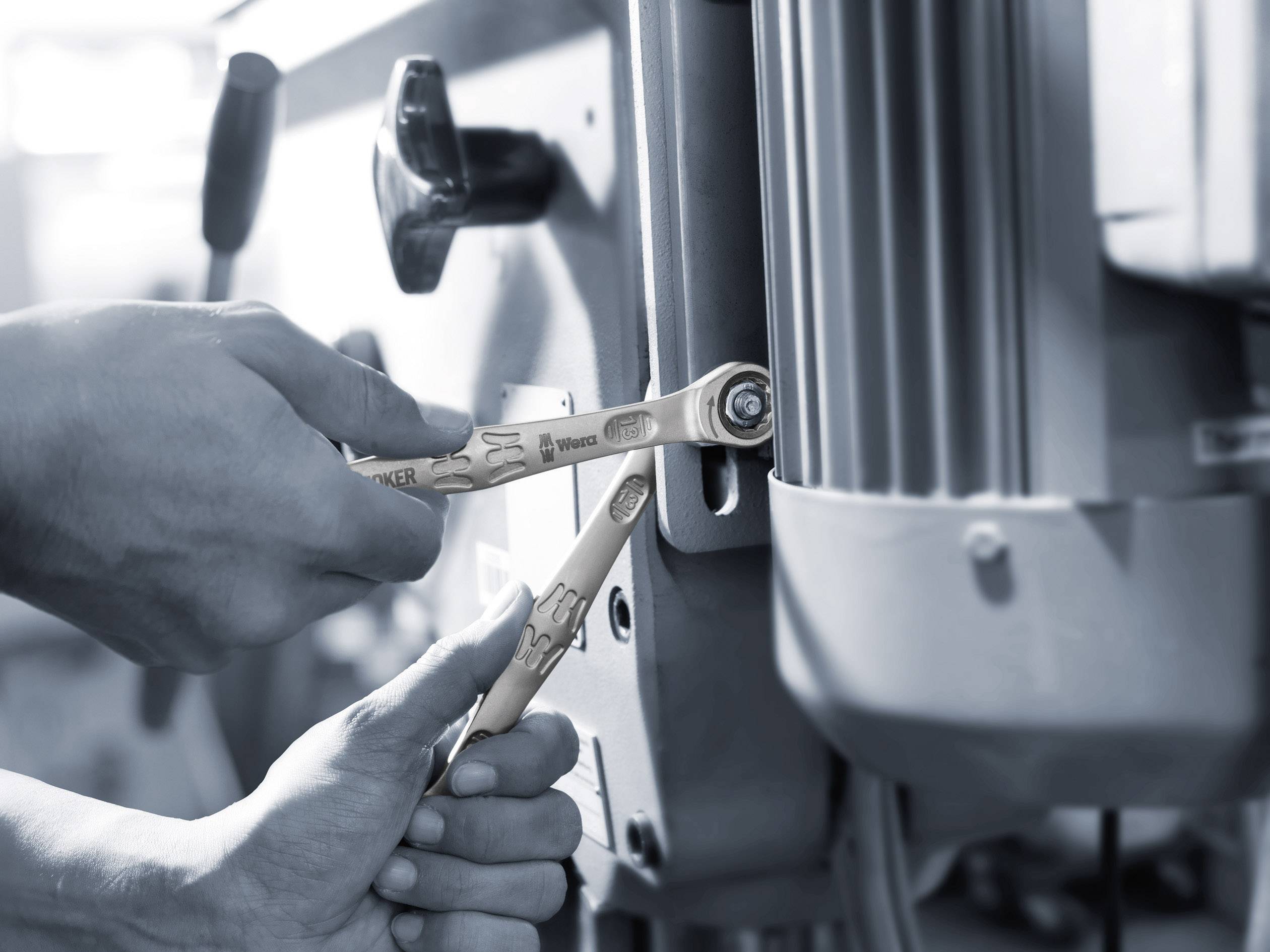 Zwei Hände benutzen Schraubenschlüssel, um eine Maschine zu reparieren. Der Fokus liegt auf der präzisen Befestigung von Schrauben.