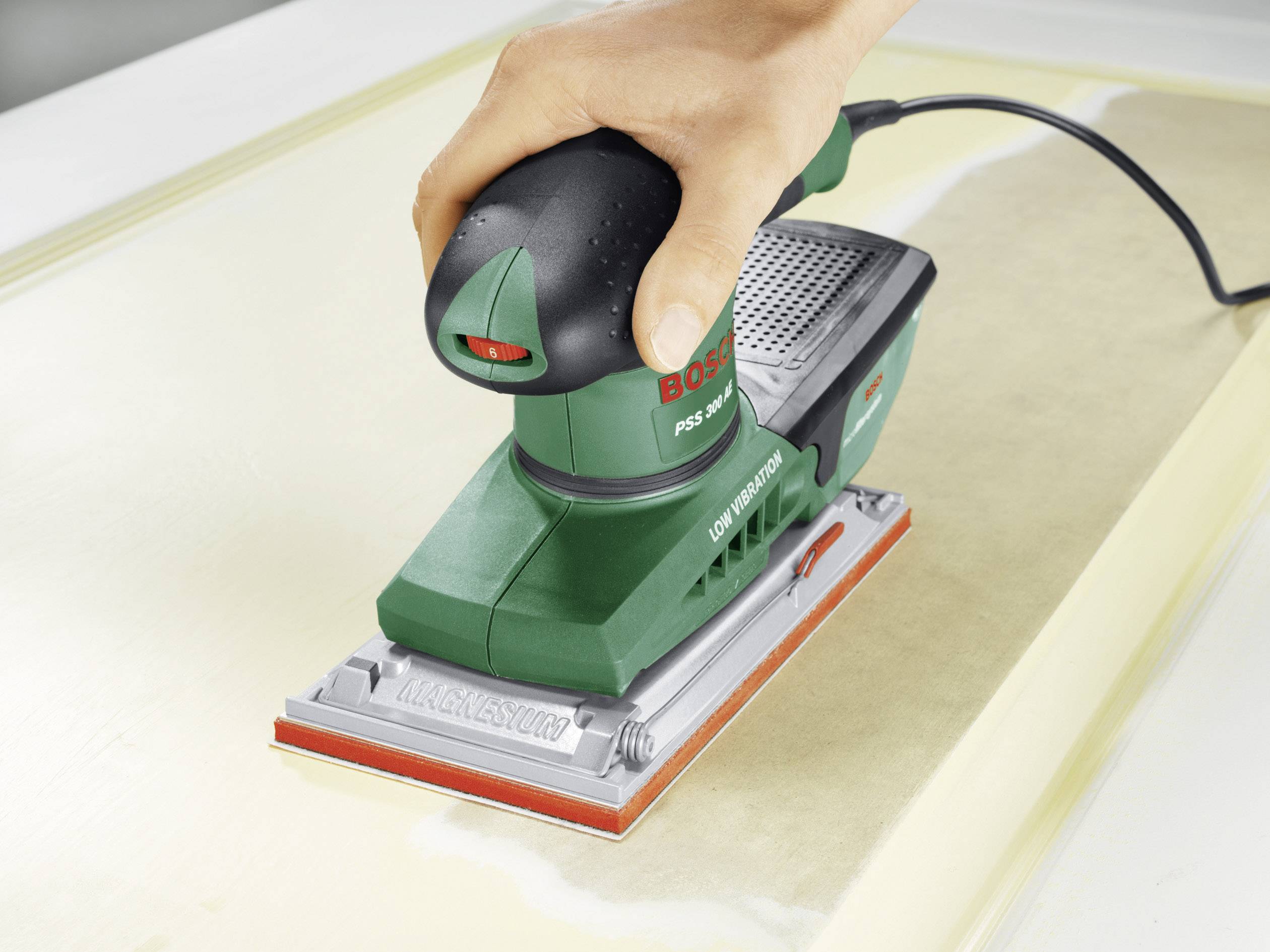 Eine Hand hält einen grünen Bosch-Schleifer, der auf einer Holzoberfläche arbeitet. Der Schleifer ist elektrisch und mit einem Kabel verbunden.