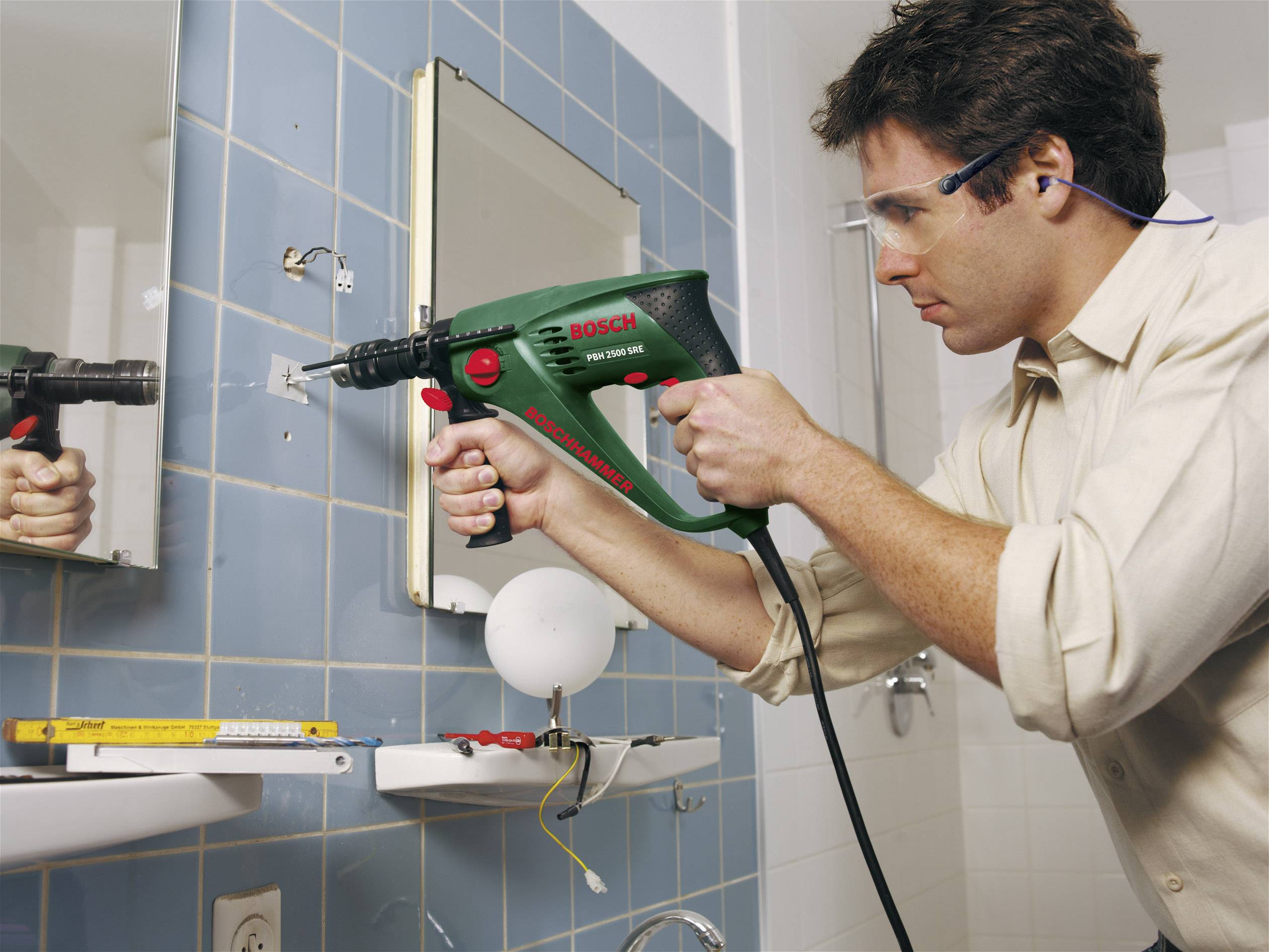 Ein Mann mit Schutzbrille benutzt eine Bohrmaschine, um ein Loch in eine geflieste Badezimmerwand zu bohren. Werkzeug liegt in der Nähe.