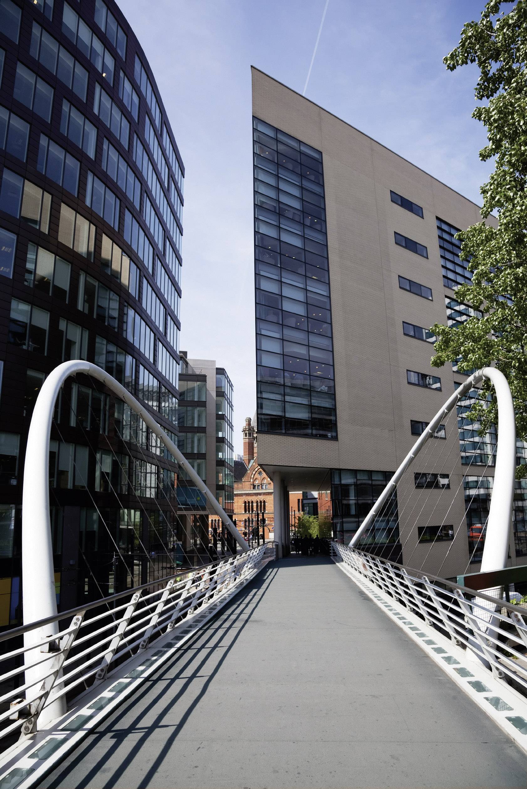 Eine moderne Fußgängerbrücke führt zwischen zwei hohe Bürogebäude. Im Hintergrund steht ein kleineres, älteres Gebäude aus rotem Backstein.