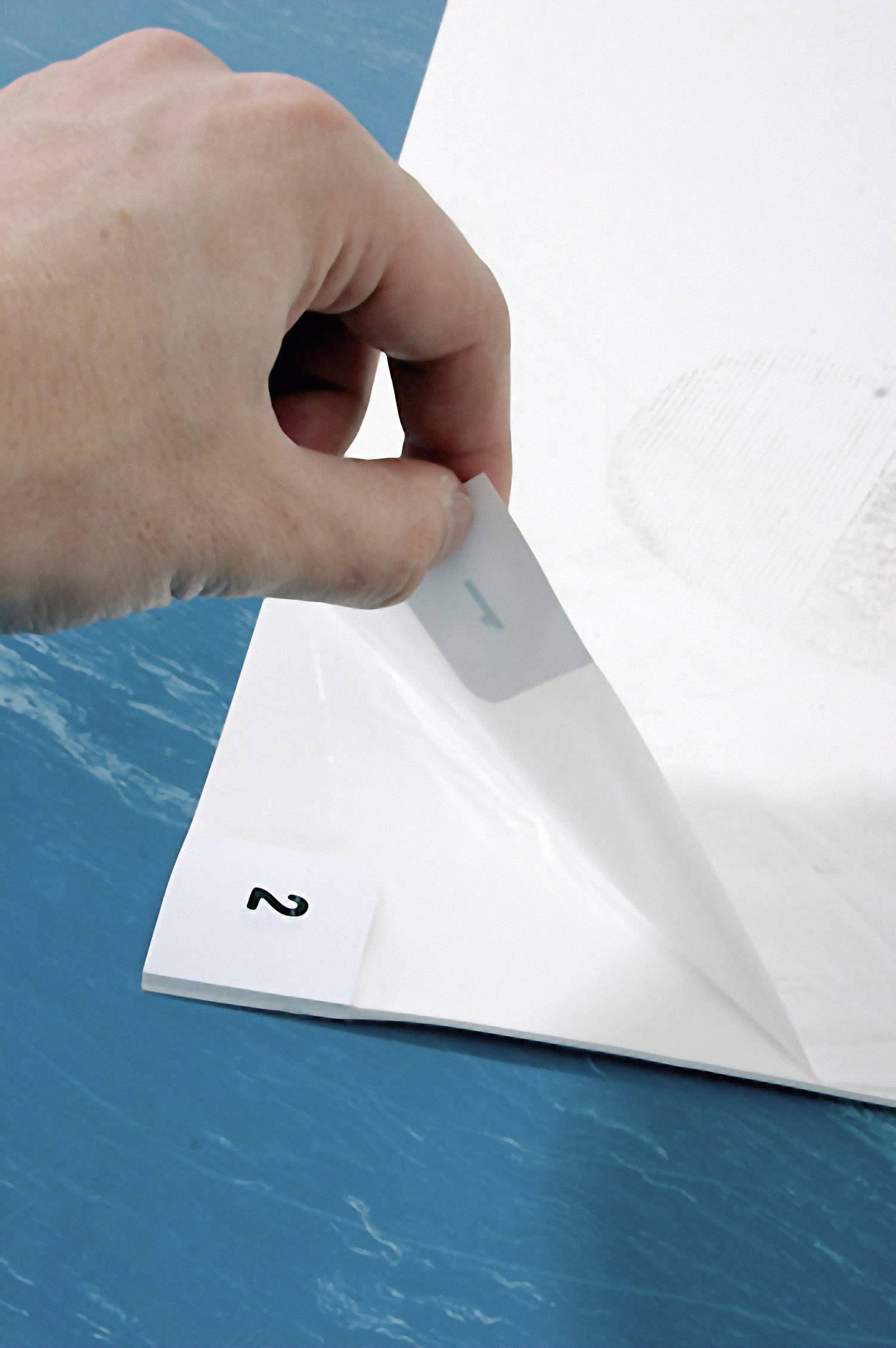 Hand hebt eine transparente Folie von einem Blatt Papier mit der Zahl '2' am Rand ab, auf blauem Hintergrund.