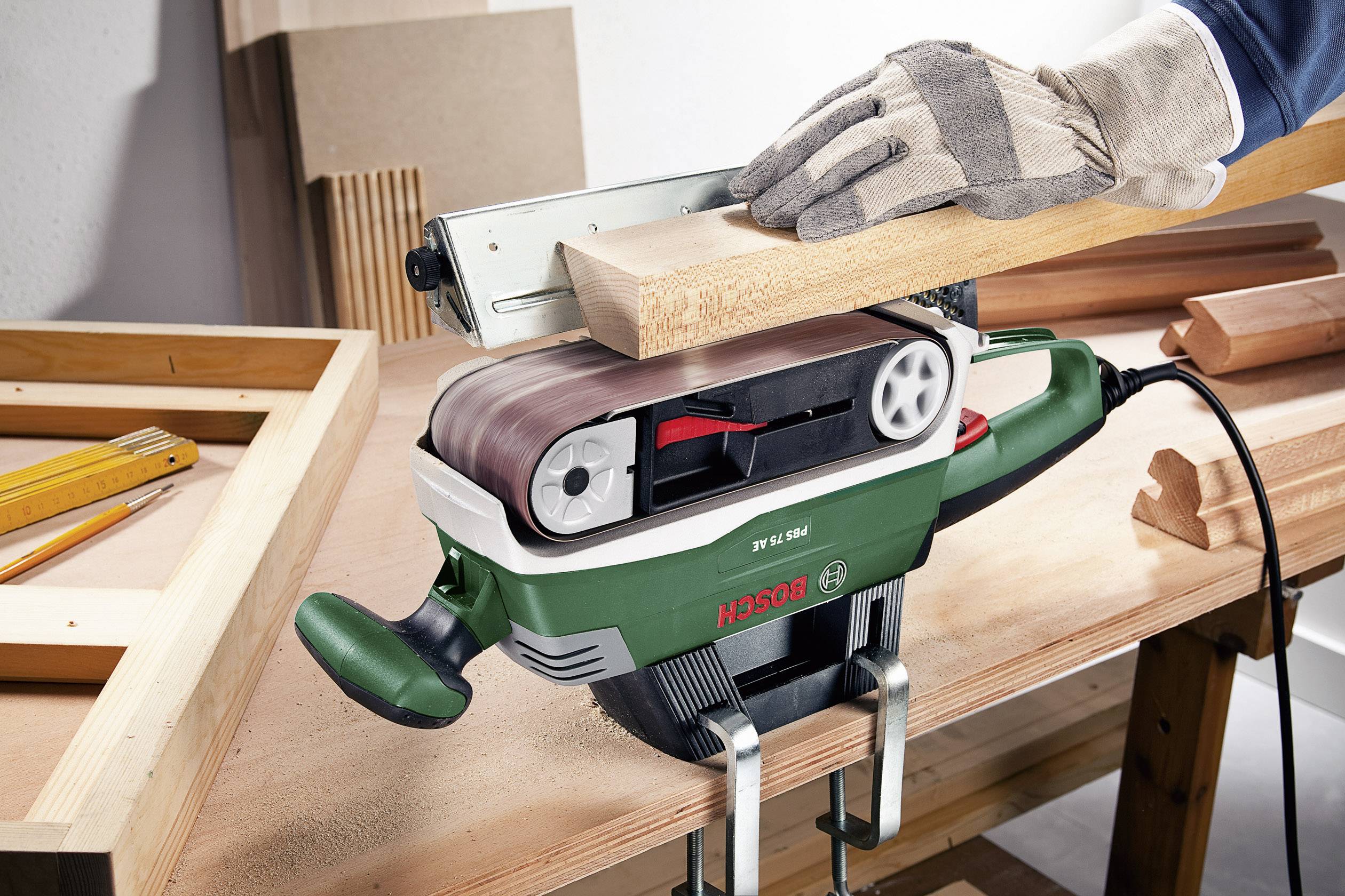 Eine Hand hält ein Stück Holz, das mit einem Bandschleifer bearbeitet wird. Der Schleifer steht auf einem Tisch in einer Werkstatt.