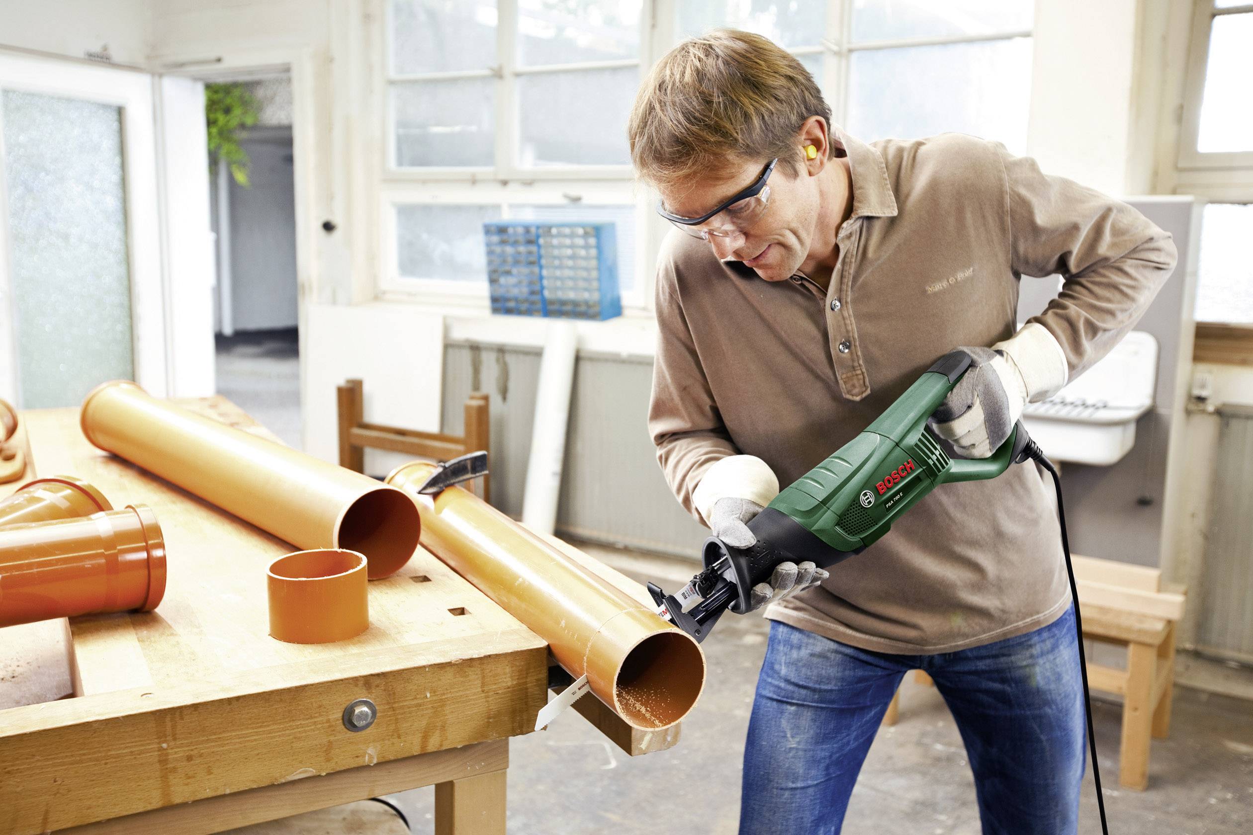 Ein Mann in Schutzbrille und Gehörschutz schneidet mit einer elektrischen Säge ein orangefarbenes Kunststoffrohr in einer Werkstatt.