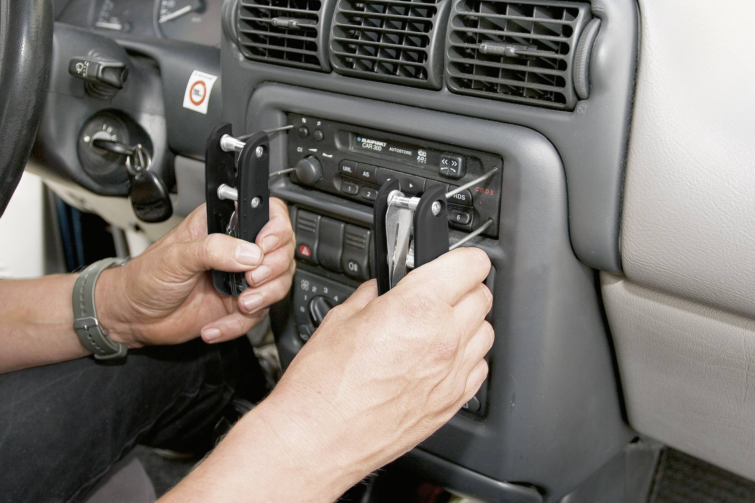 Eine Person entfernt mit Werkzeug ein Autoradio aus einem Fahrzeugarmaturenbrett. Hände und Bedienelemente sichtbar.