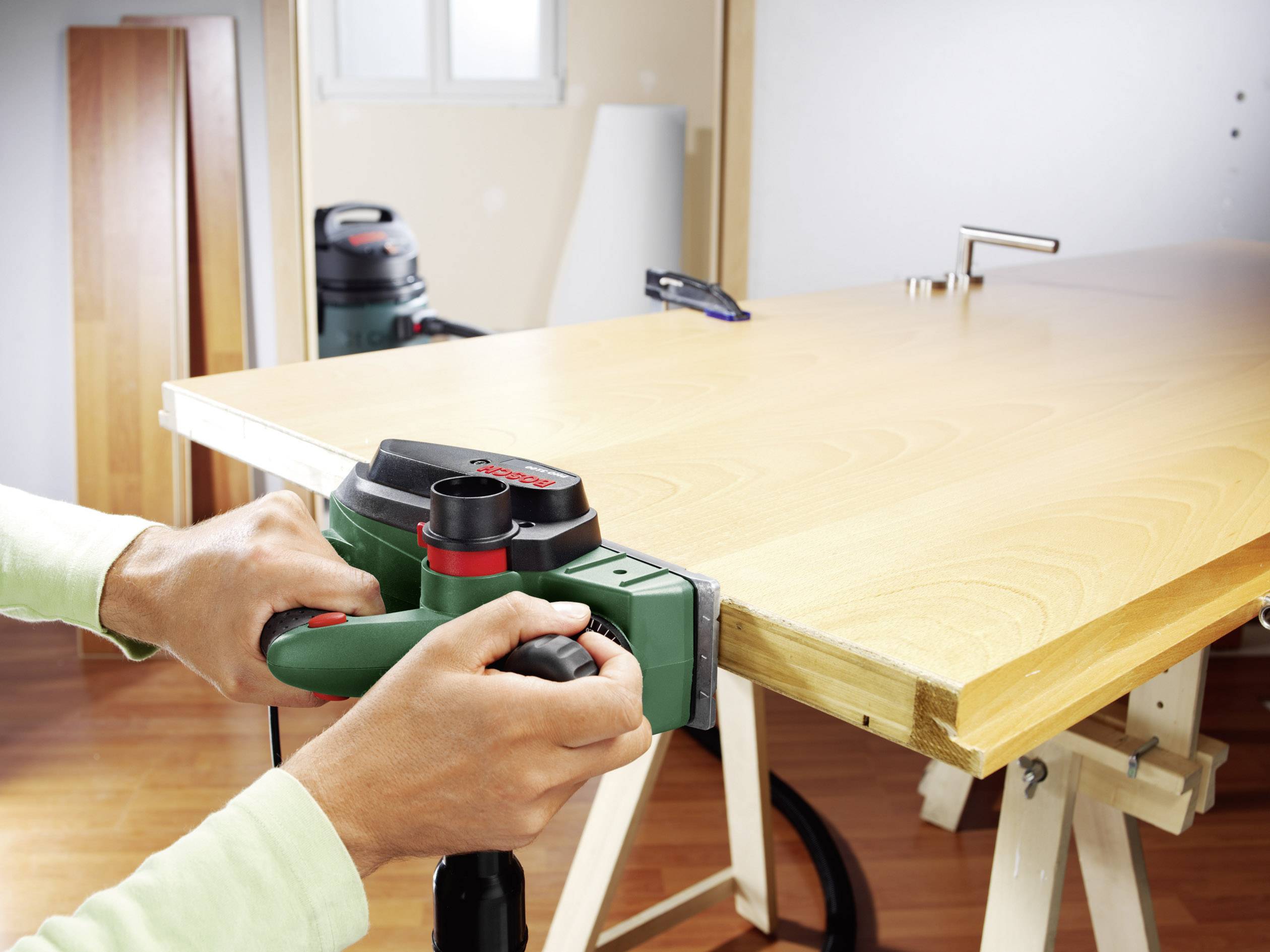Eine Person führt Holzarbeiten auf einem Tisch durch. Sie verwendet ein elektrisches Werkzeug, um die Tischkante zu glätten. Im Hintergrund befindet sich eine Werkstattumgebung.