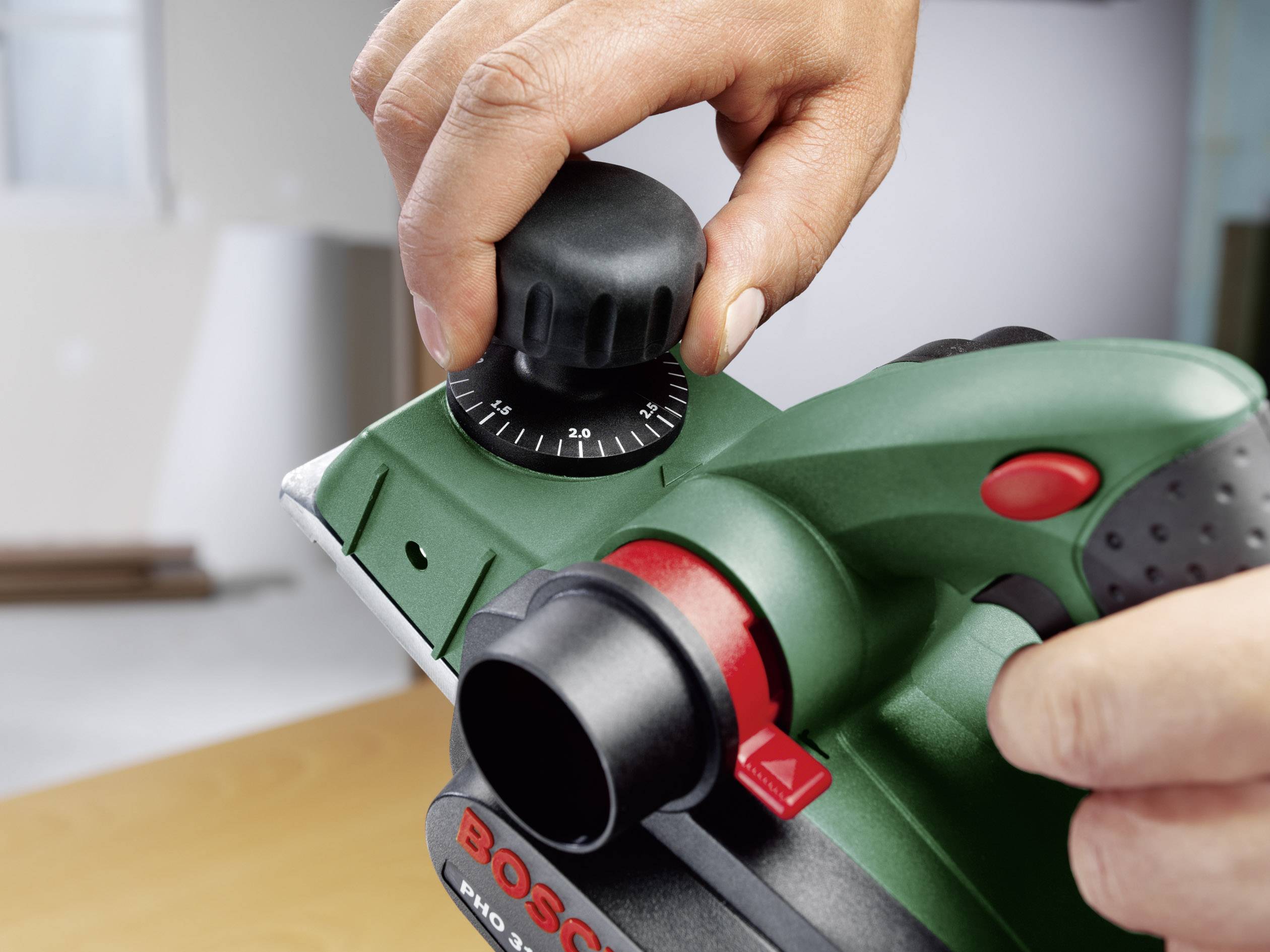 Person stellt einen grünen Elektrohobel ein, indem sie am Einstellknopf dreht. Gerät ist auf einer Tischplatte positioniert.