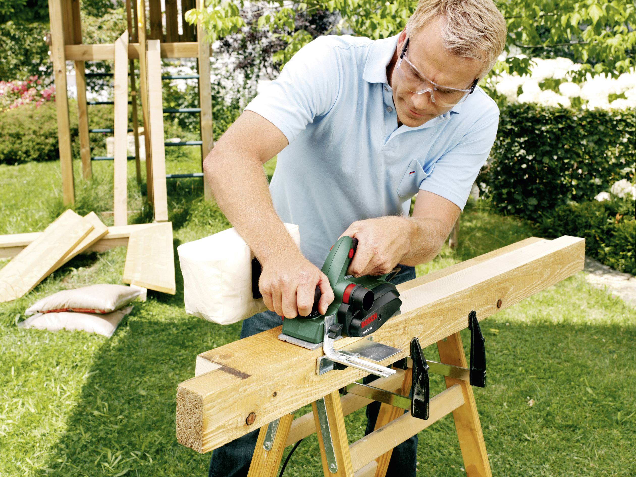 Ein Mann im Garten arbeitet mit einer Bandsäge an einem Holzbrett, geschützt durch eine Schutzbrille.