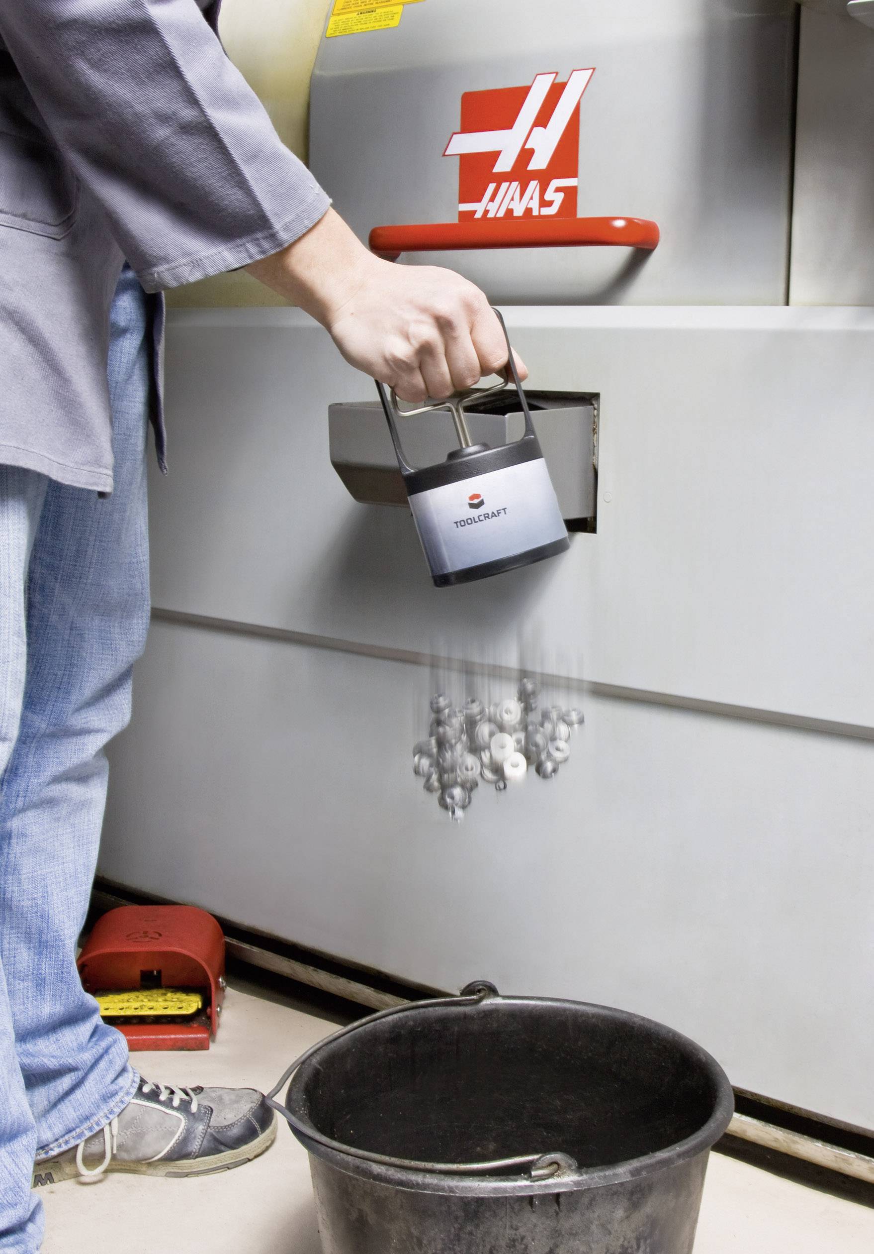 Person entleert Kanne mit Metallteilen in schwarzen Eimer vor industrieller Maschine, gekennzeichnet mit rotem 'Haas'-Logo.