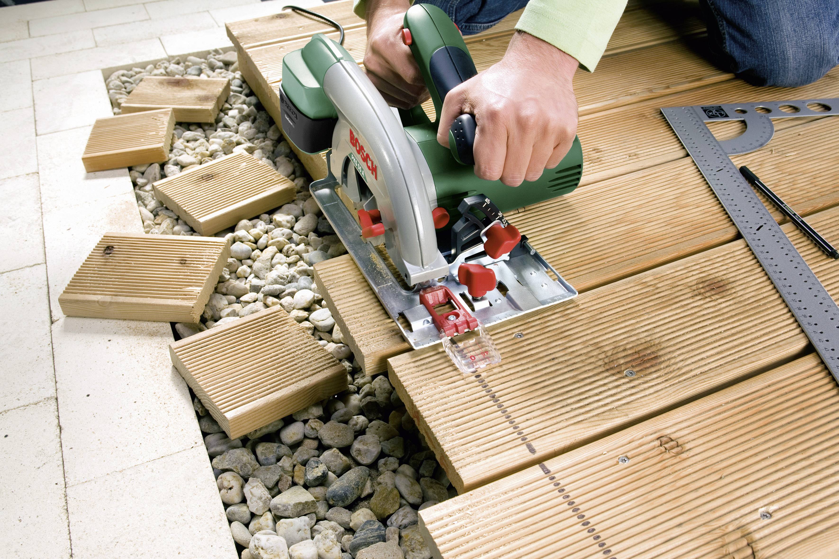 Eine Person sägt Holz mit einer Kreissäge auf einer Terrasse. Holzspäne und Kies im Hintergrund verdeutlichen den Heimwerkerkontext.