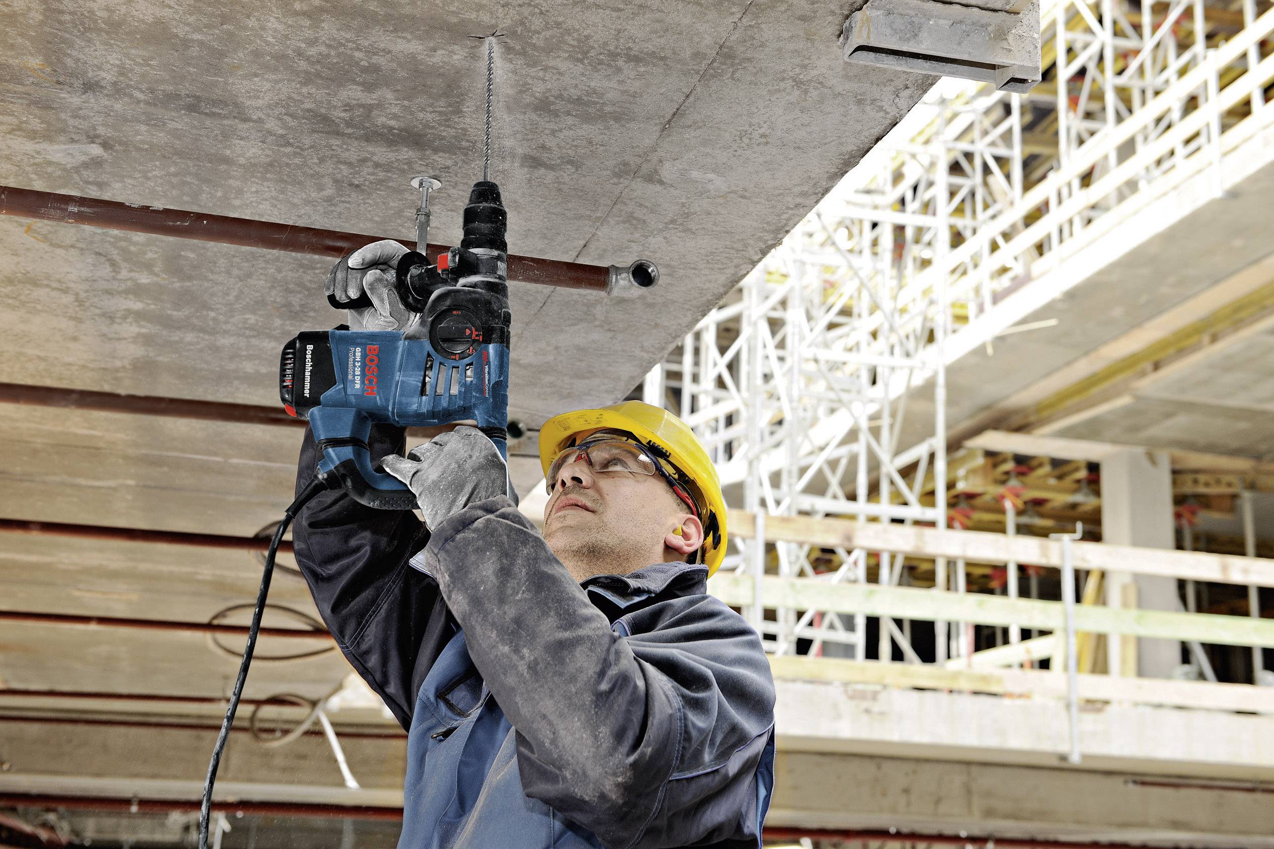 Ein Bauarbeiter trägt einen gelben Schutzhelm und benutzt eine Bohrmaschine, um in eine Betondecke auf einer Baustelle zu bohren.