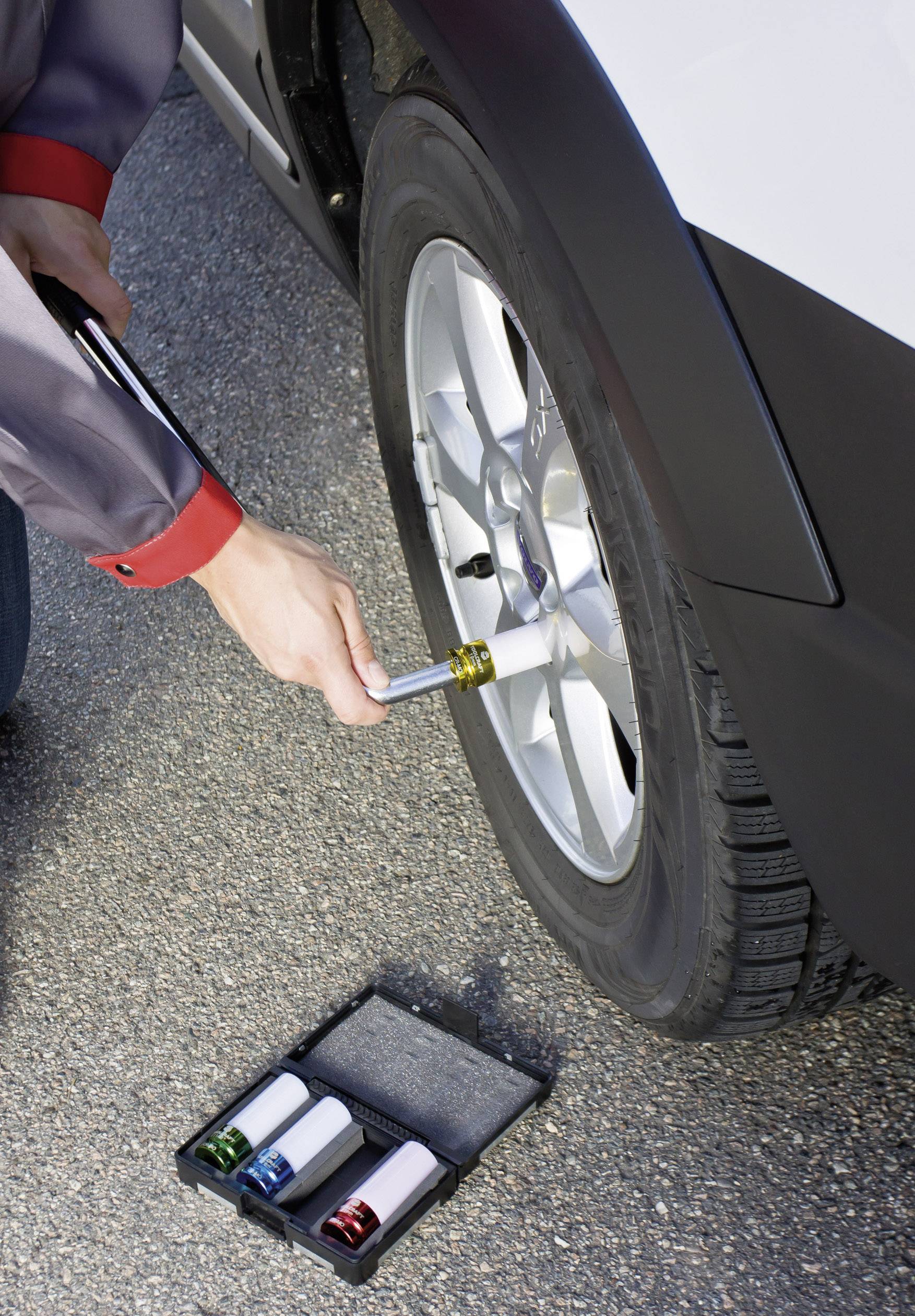 Person wechselt Autoreifen mit Schraubenschlüssel, Nahaufnahme. Werkzeugkasten am Boden mit bunten Stecknüssen.