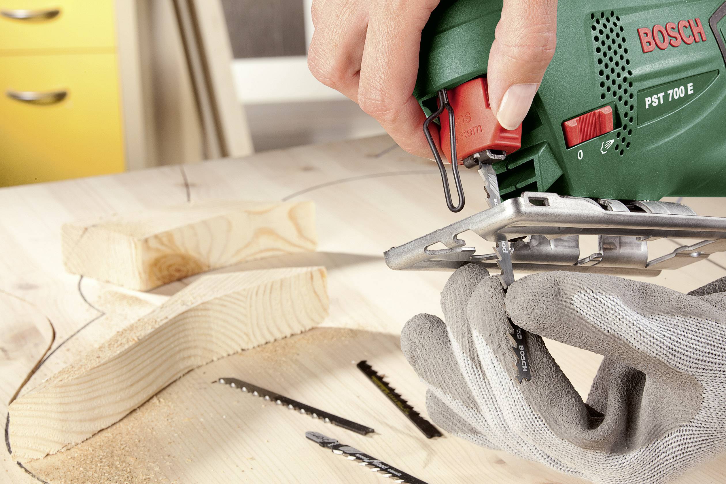 Eine Person sägt eine kurvige Form aus einem Holzbrett mit einer grünen Stichsäge, während sie graue Schutzhandschuhe trägt.