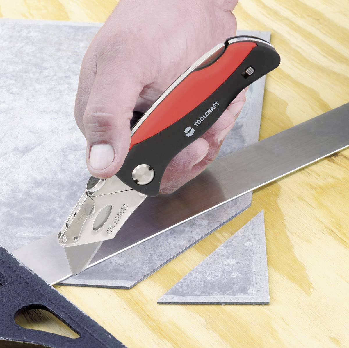 Eine Hand benutzt ein Universalmesser, um ein dünnes Blech entlang eines Lineals auf einem Holztisch zu schneiden.