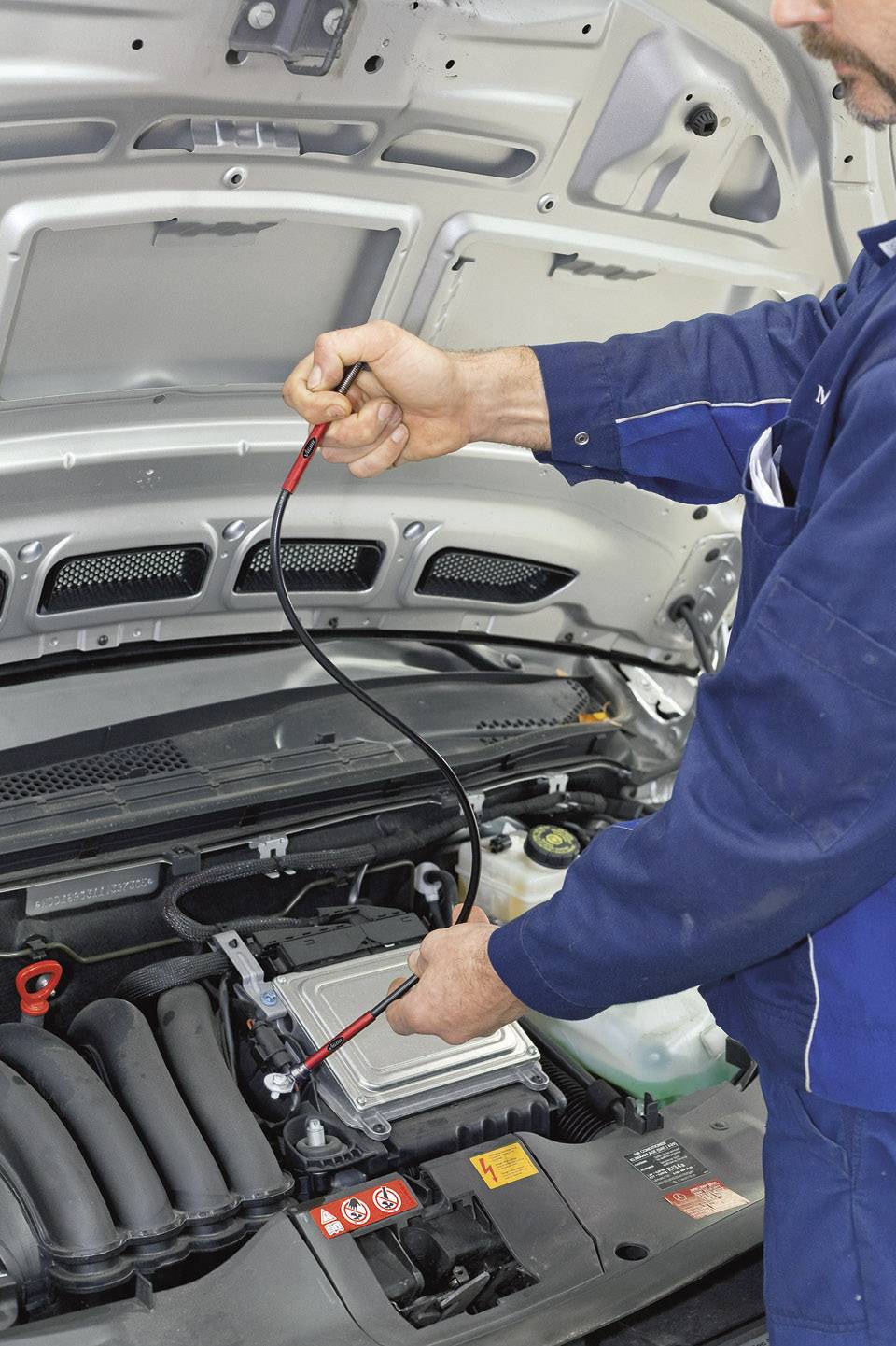 Automechaniker in blauer Uniform prüft den Motor eines Autos mit einem Diagnosetool unter geöffneter Motorhaube.