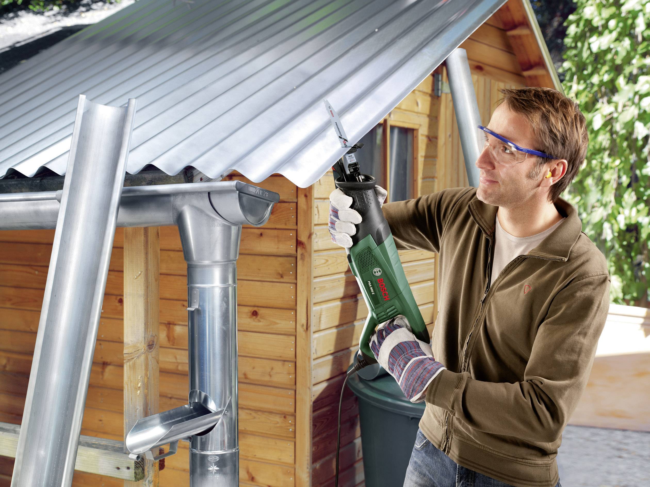 Ein Mann arbeitet mit einer Säbelsäge an einem Wellblechdach neben einem Holzschuppen. Er trägt Schutzbrille und Handschuhe.