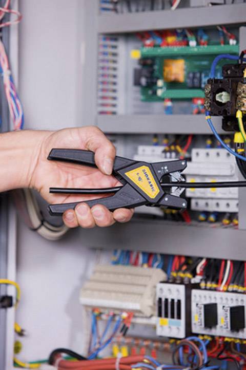 Eine Hand hält ein Crimpwerkzeug vor einem offenen Schaltschrank mit vielen Kabeln und elektrischen Komponenten.