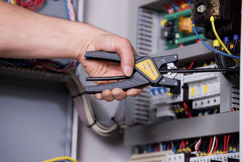 Eine Hand hält ein Werkzeug und arbeitet an einem offenen Schaltschrank mit Kabeln und elektrischen Komponenten im Inneren.