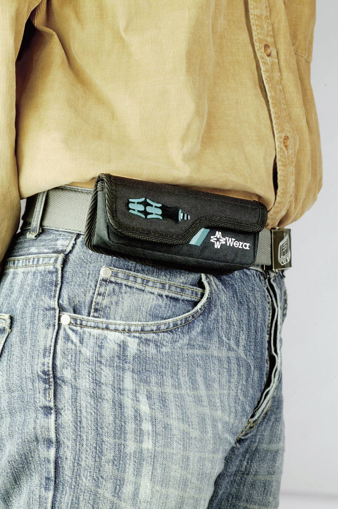Eine Person trägt eine schwarze Werkzeugtasche der Marke Wera an einem grauen Gürtel. Die Person trägt blaue Jeans und ein beiges Hemd.