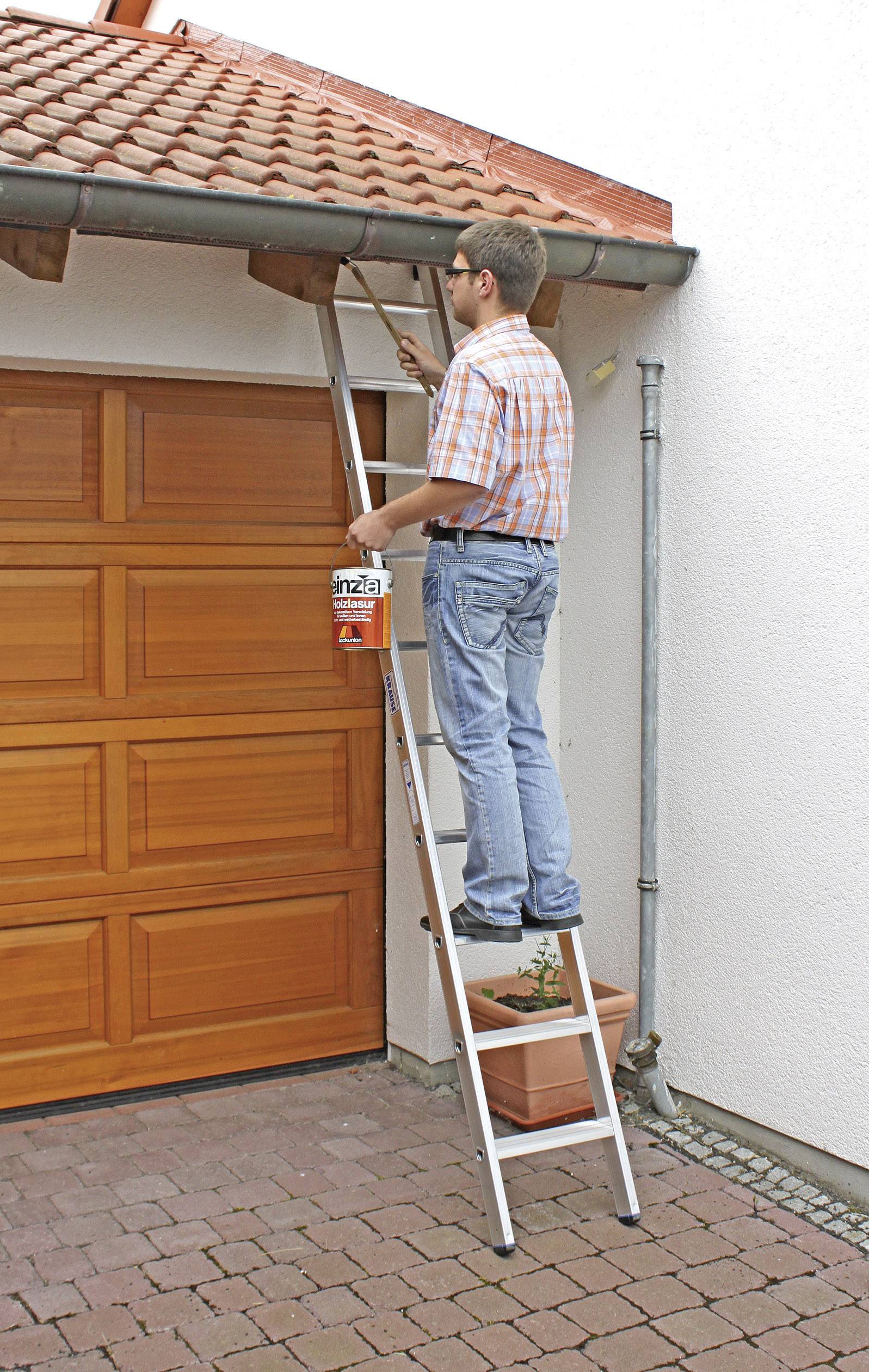 Eine Person steht auf einer Leiter und streicht die Dachrinne an einem Haus. Links ist ein Holztor zu sehen.