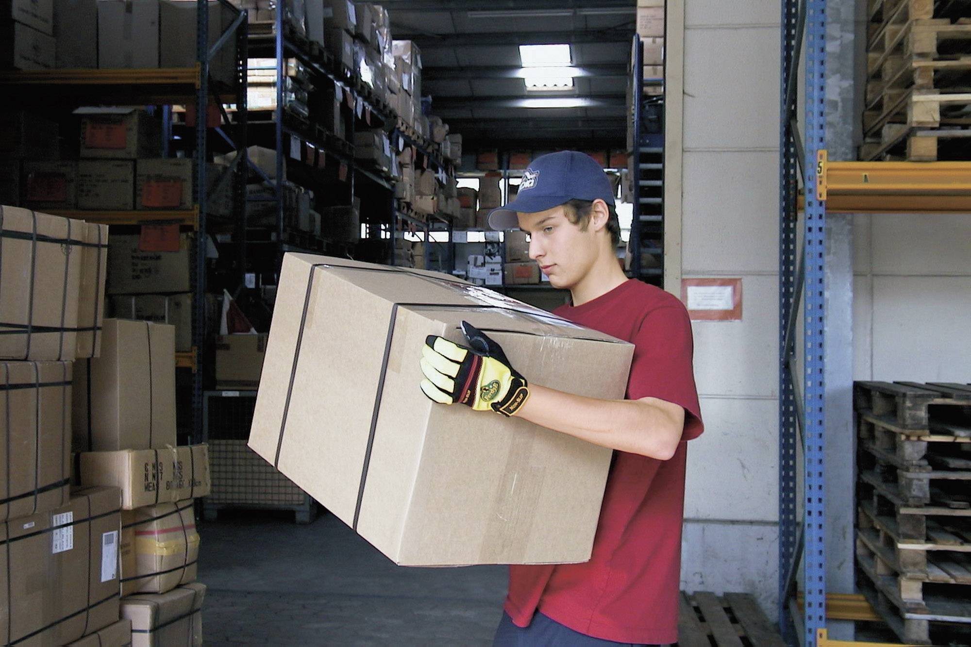Ein Mann in rotem T-Shirt und blauen Hut trägt ein großes, braunes Paket in einem Lagerhaus mit vielen Regalen im Hintergrund.