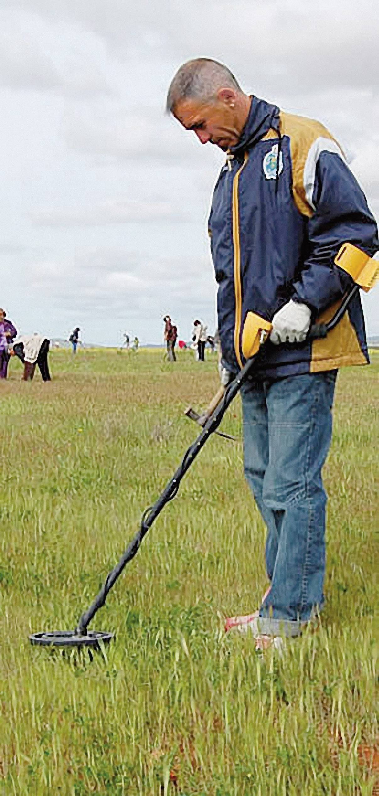Ein Mann mit Metalldetektor sucht in einem Feld nach Metallgegenständen. Im Hintergrund sind unscharf andere Personen zu sehen.