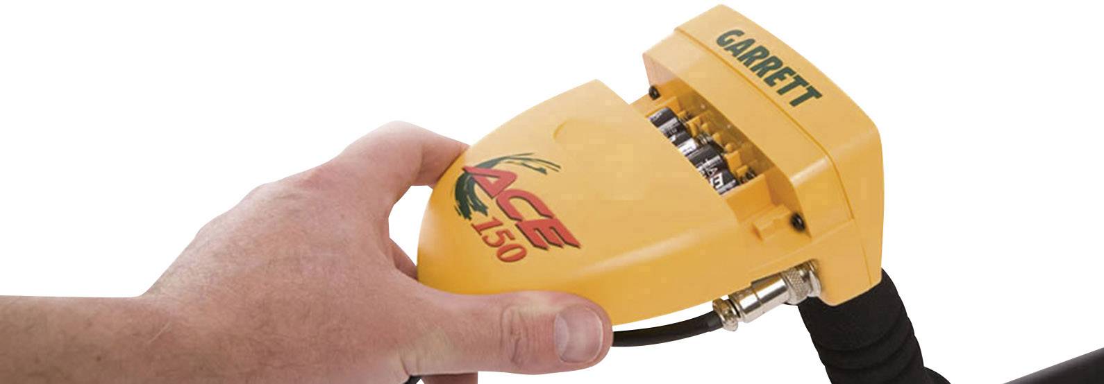 Hand hält gelbes Gerät mit der Aufschrift 'ACE 150', verwendet zur Metallsuche. Oben steht 'GARRETT'. Kabel ist angeschlossen.