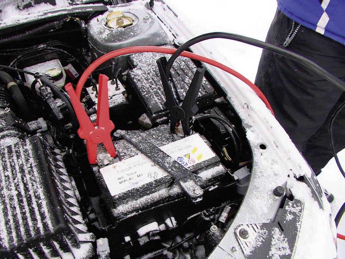 Autobatterie wird mit Starthilfekabeln überbrückt, rote und schwarze Klemme angeschlossen. Winterliche Umgebung, Schnee sichtbar.