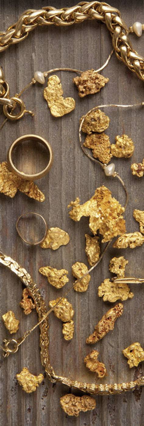 Goldnuggets und Schmuckstücke liegen auf einer rustikalen, braunen Holzoberfläche verteilt, darunter Ringe und Halsketten in Goldtönen.