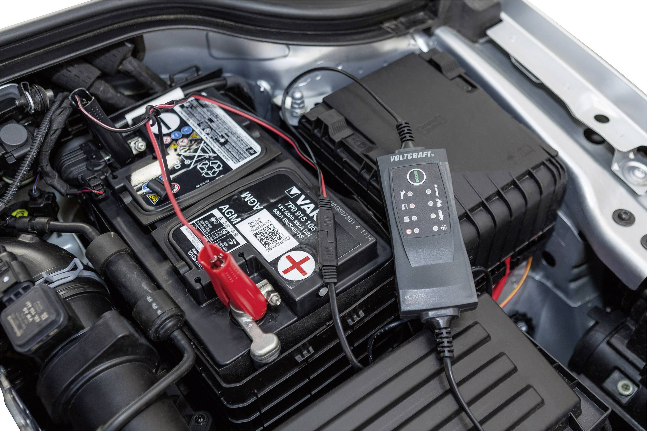 Autobatterie im Motorraum mit angeschlossenem Ladegerät. Das Ladegerät zeigt den Ladezustand an und versorgt die Batterie mit Strom.