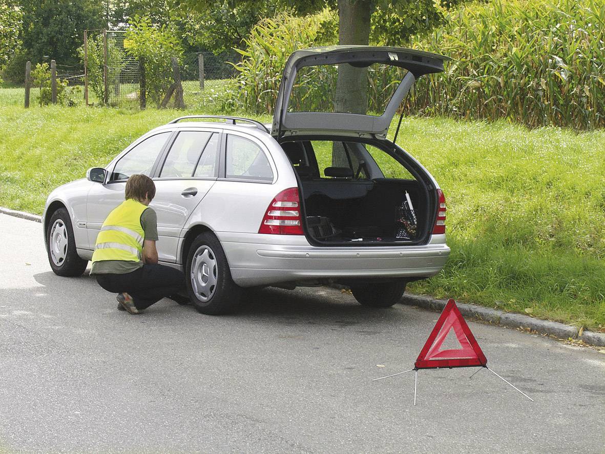 Ein silbernes Auto mit offenem Kofferraum steht am Straßenrand. Eine Person mit Warnweste kniet neben dem Auto. Ein Warndreieck ist aufgestellt.
