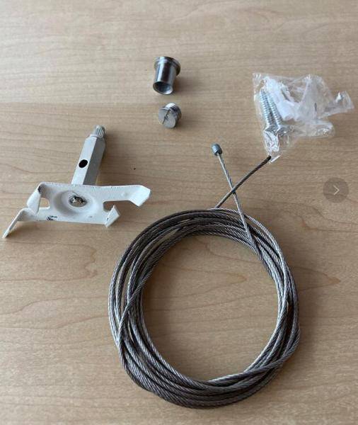 Set von Metallteilen und Drahtseil auf Holztisch. Enthält Halterung, Gewinde und Schrauben, vermutlich für eine Befestigungslösung.