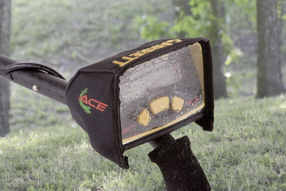 Ein Metall-Detektor mit einer regennassen Abdeckung zeigt ein Display an. Der Hintergrund ist eine grüne, bewaldete Landschaft.