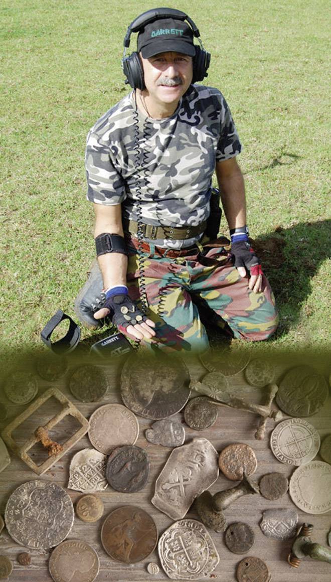 Eine Person kniet auf Gras mit Metalldetektor, trägt Tarnkleidung. Im Vordergrund eine Sammlung von gefundenen Münzen und Artefakten.