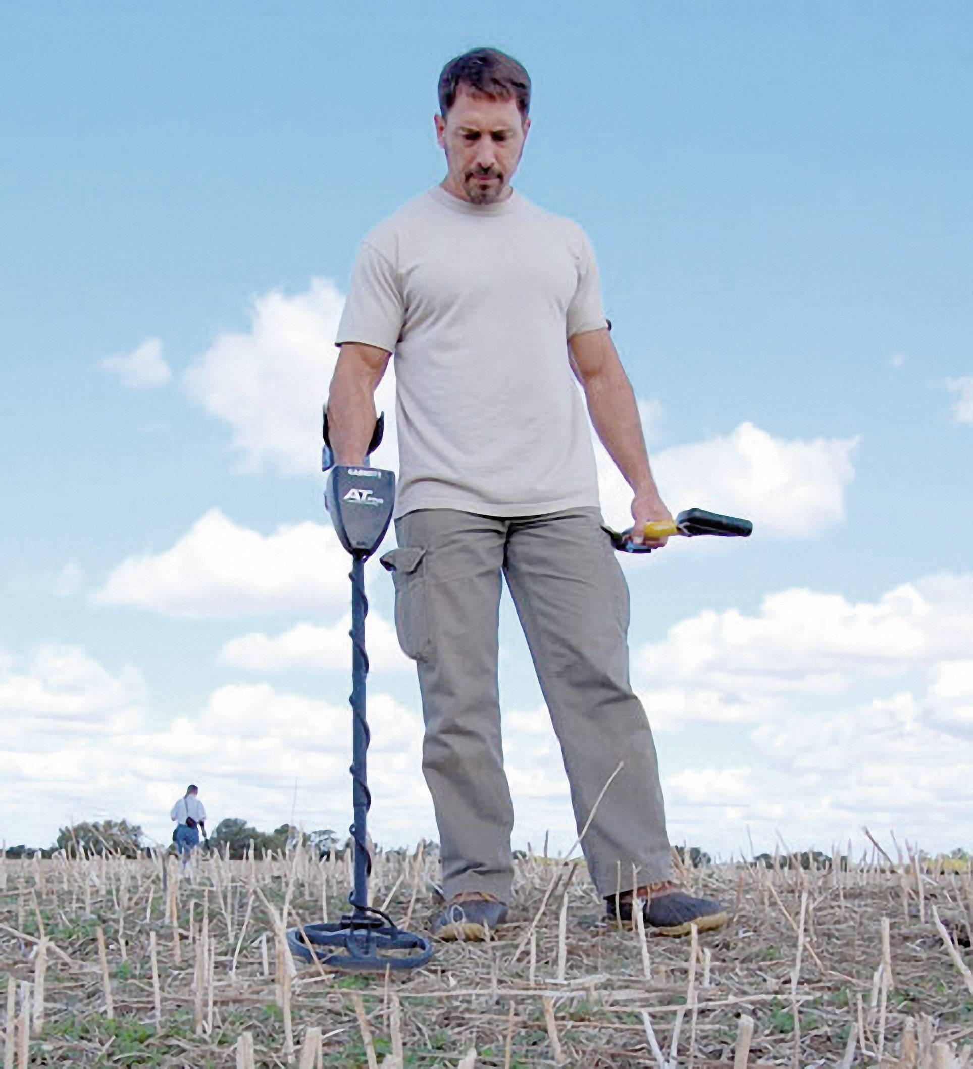 Ein Mann sucht mit einem Metalldetektor auf einem Feld, ein weiterer Mensch im Hintergrund. Der Himmel ist blau mit einigen Wolken.