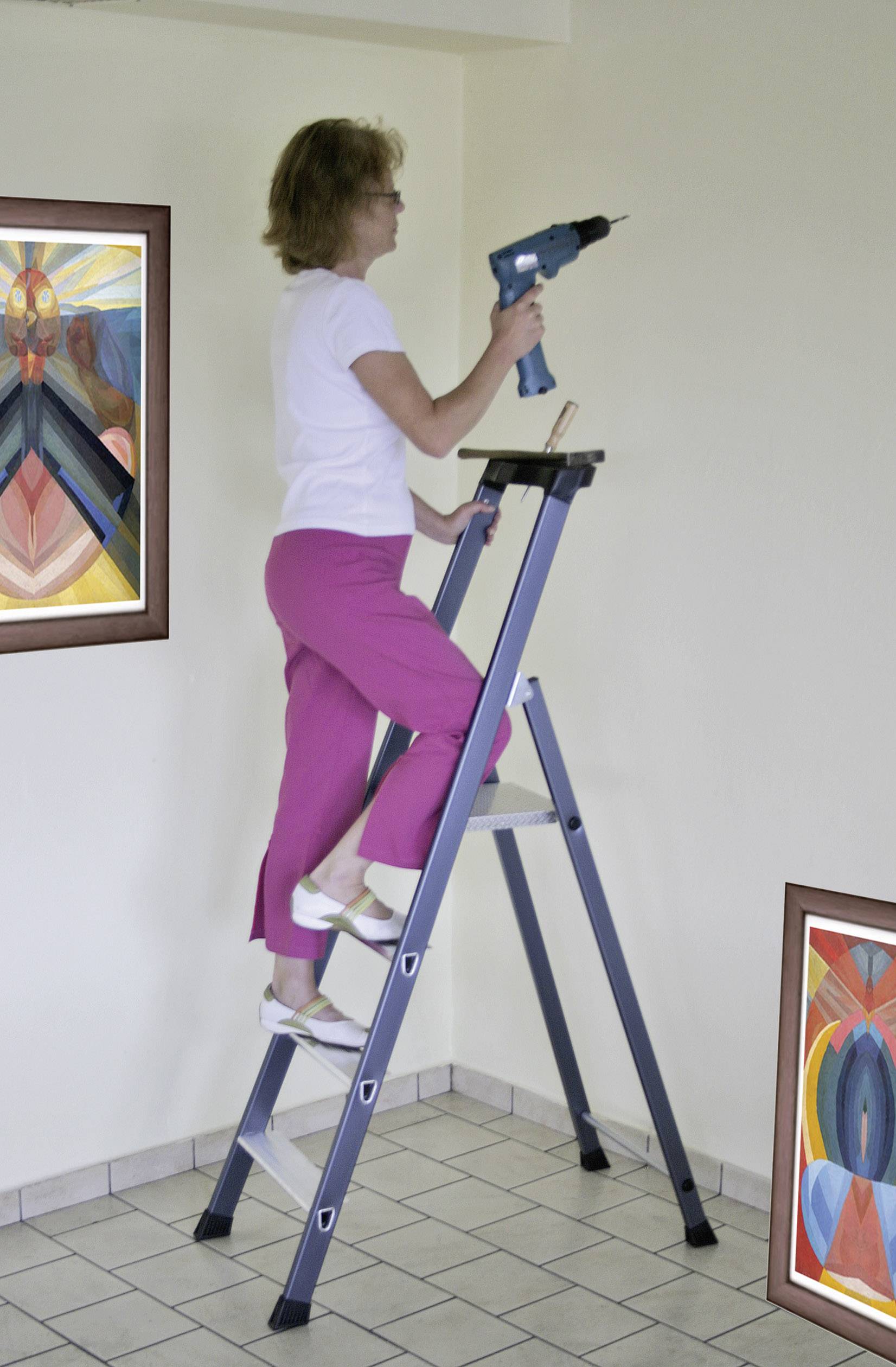 Eine Frau steht auf einer Leiter und hält einen Bohrer, um ein Bild an der Wand aufzuhängen. Zwei Gemälde sind bereits aufgehängt.