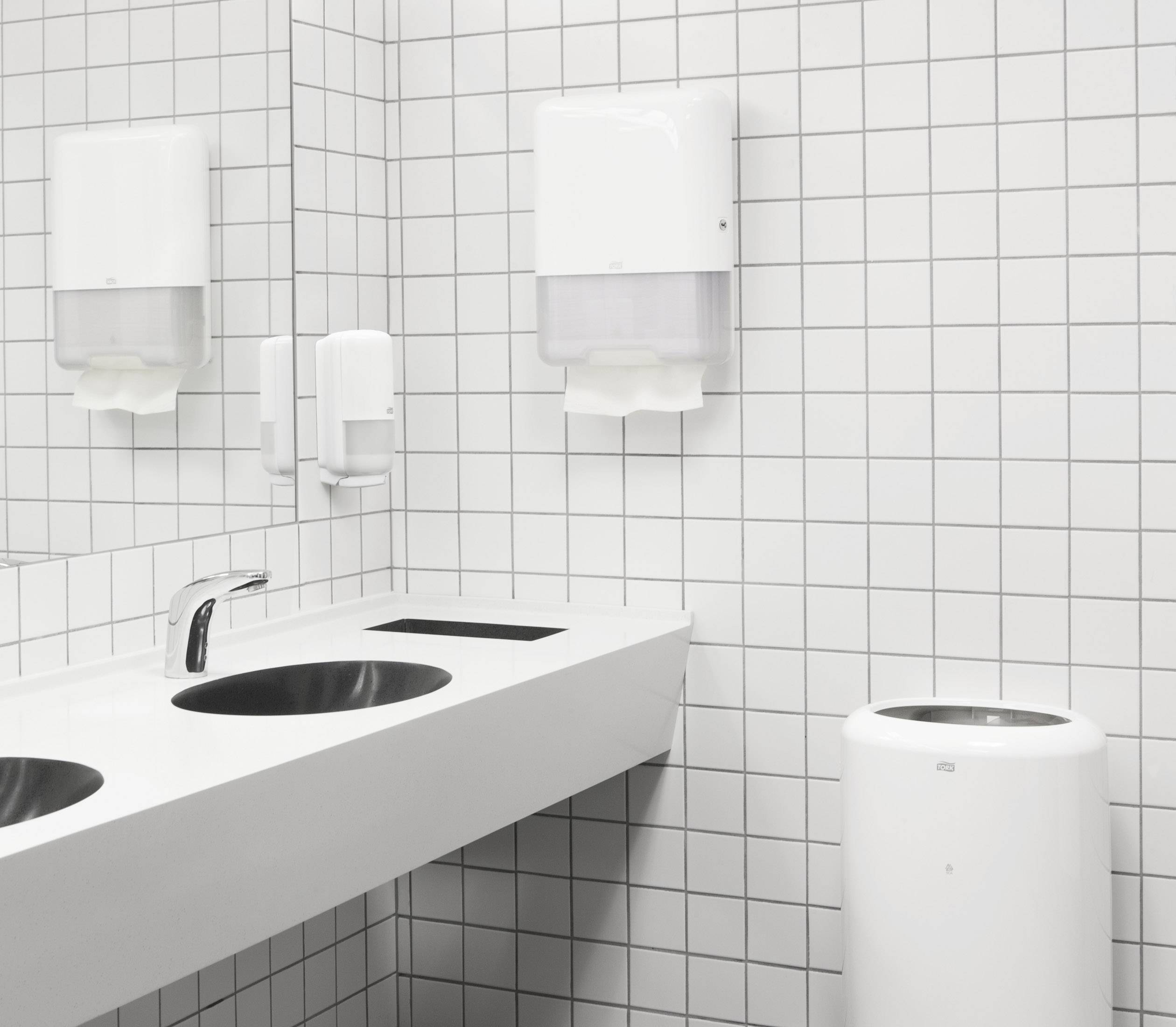 Öffentliche Toiletten mit weißen, gefliesten Wänden, zwei Waschbecken und einem Spender für Seife oder Papierhandtücher.