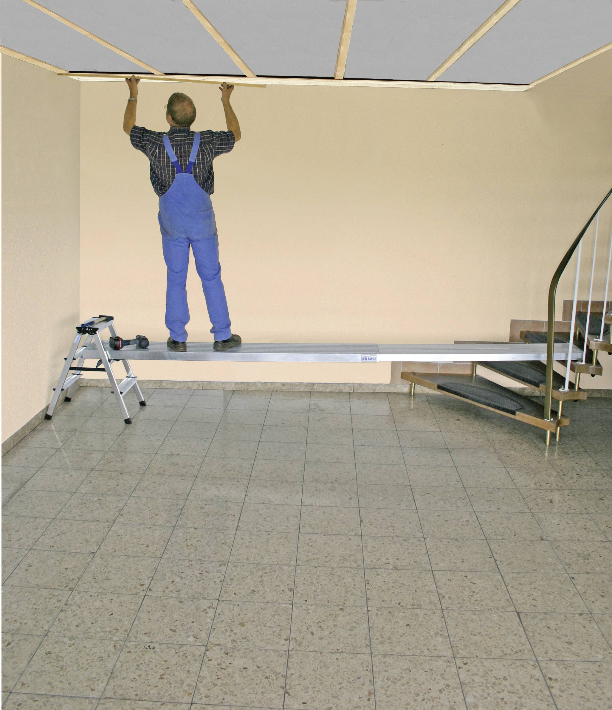 Ein Handwerker in blauer Latzhose installiert eine Deckenverkleidung in einem Raum mit Wendeltreppe und Fliesenboden.
