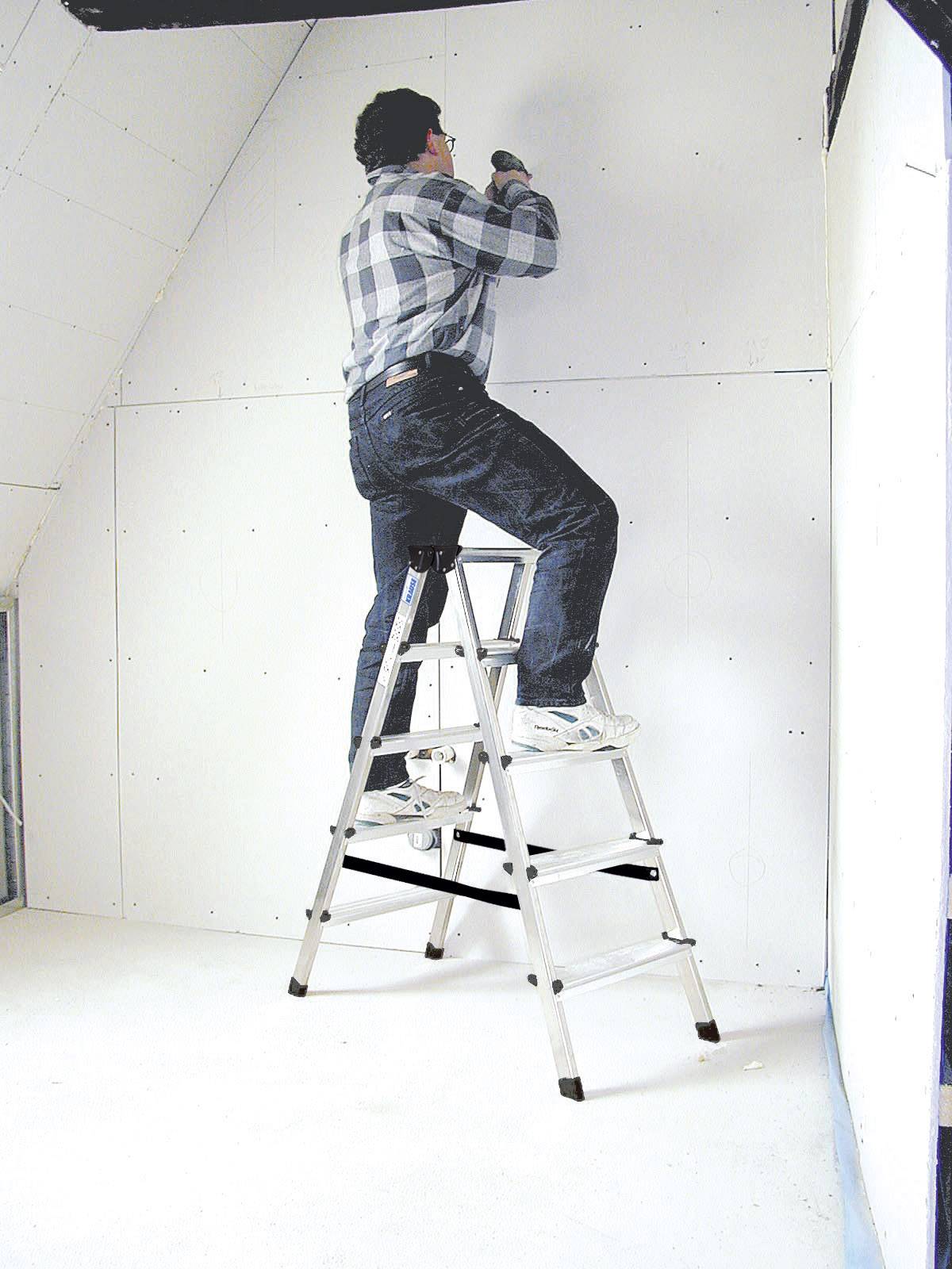 Eine Person in kariertem Hemd und Jeans steht auf einer Leiter und montiert eine Trockenbauwand in einem Raum.