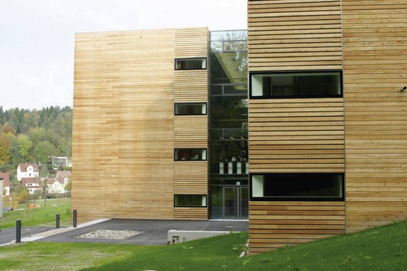 Ein modernes Gebäude mit Holzfassade und Glasfront. Im Hintergrund ist eine ländliche Umgebung mit Bäumen und Häusern sichtbar.