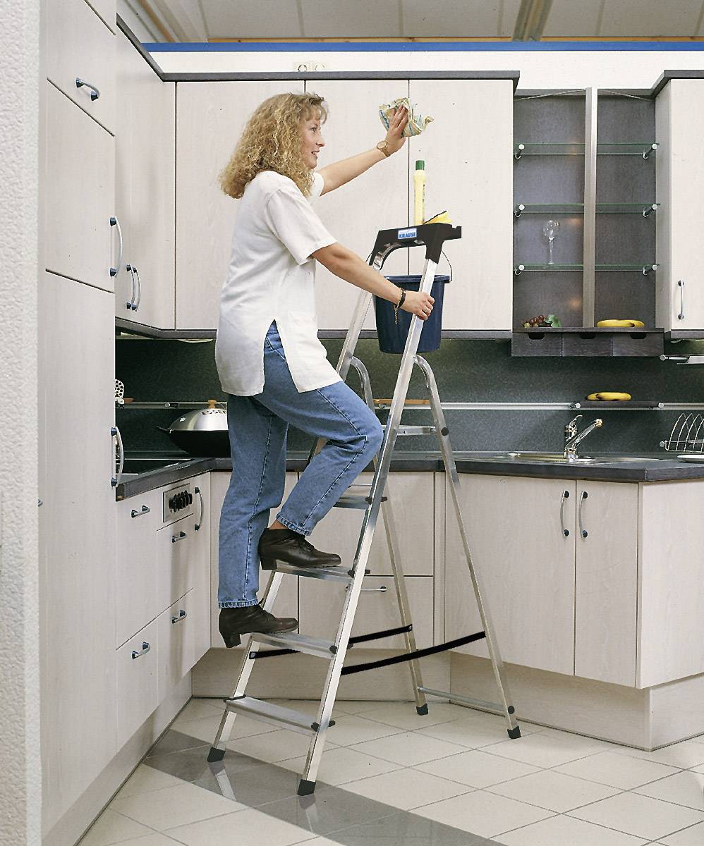 Frau auf Leiter wischt Küchenoberschränke; trägt Freizeitkleidung; moderne Küche mit hellem Holz und dunklen Arbeitsflächen.