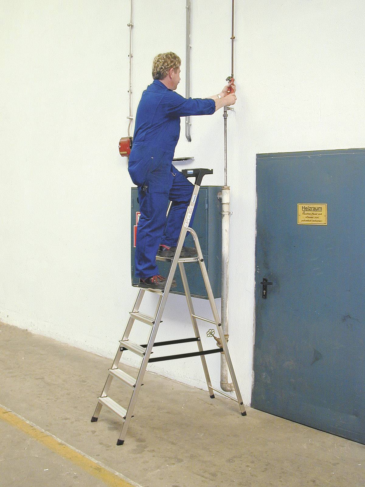 Eine Person in blauer Arbeitskleidung steht auf einer Leiter und führt Reparaturen an einer Rohrleitung an der Wand durch.