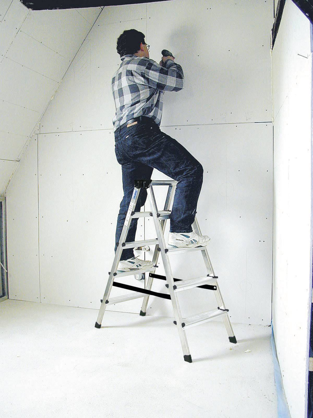 Person befestigt Trockenbauplatten an einer Wand, steht auf einer Leiter in einem unfertigen Raum, trägt Karobluse und Jeans.