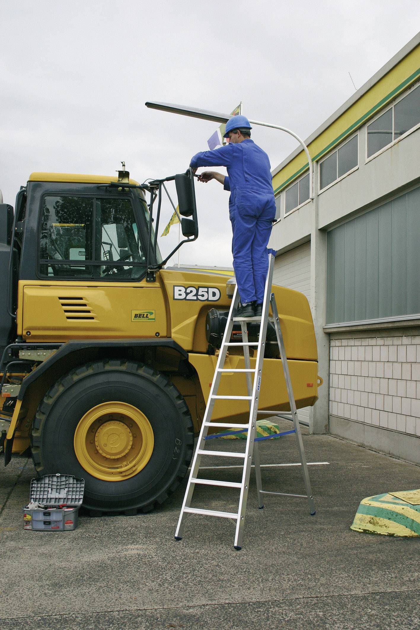 Ein Arbeiter in blauer Arbeitskleidung steht auf einer Leiter und repariert eine Straßenlaterne neben einem gelben Baufahrzeug.