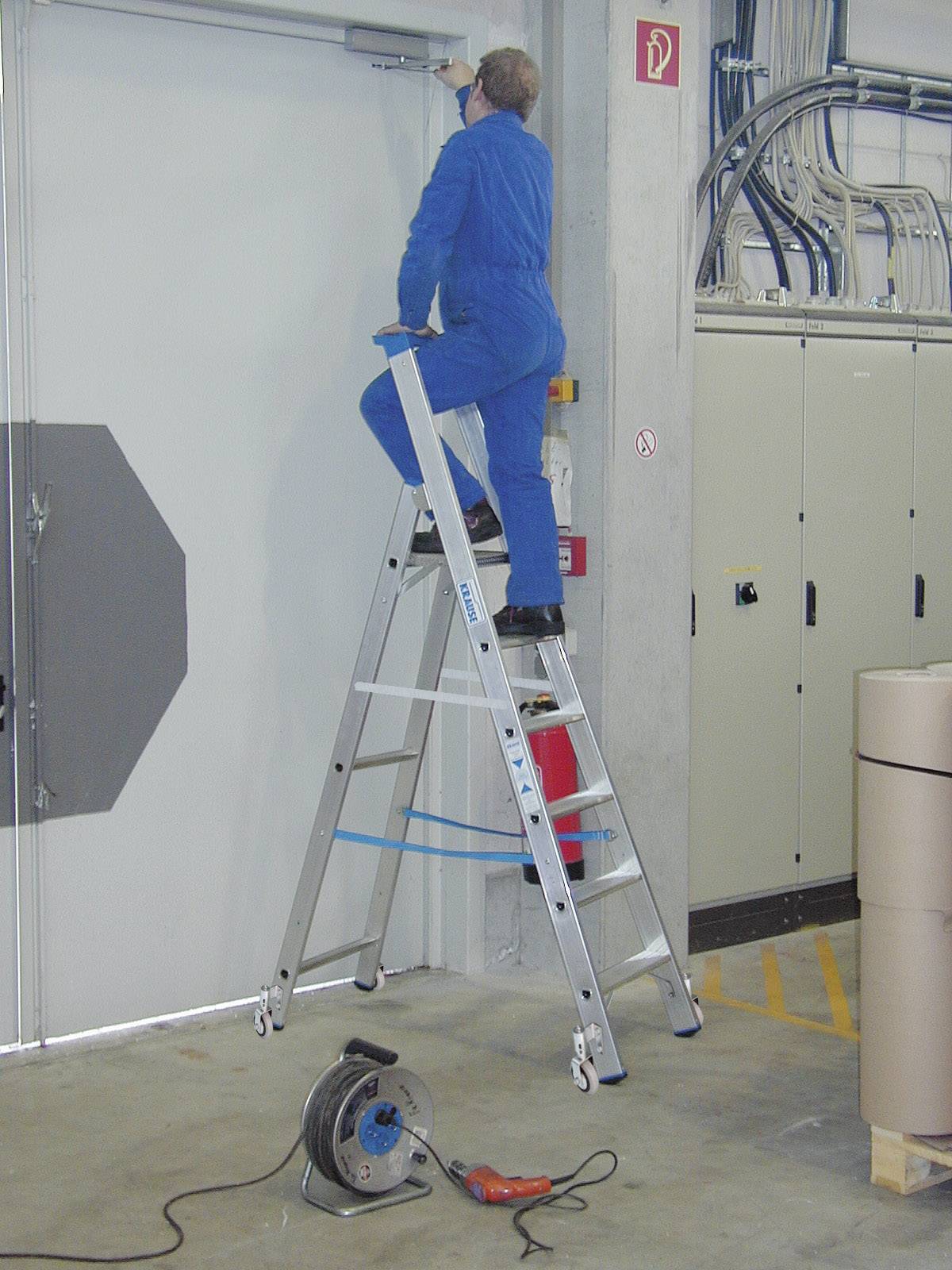 Ein Mann in blauer Arbeitskleidung steht auf einer Leiter und arbeitet an einer Überkopfarbeit in einer Werkstatt oder einem Lagerraum.