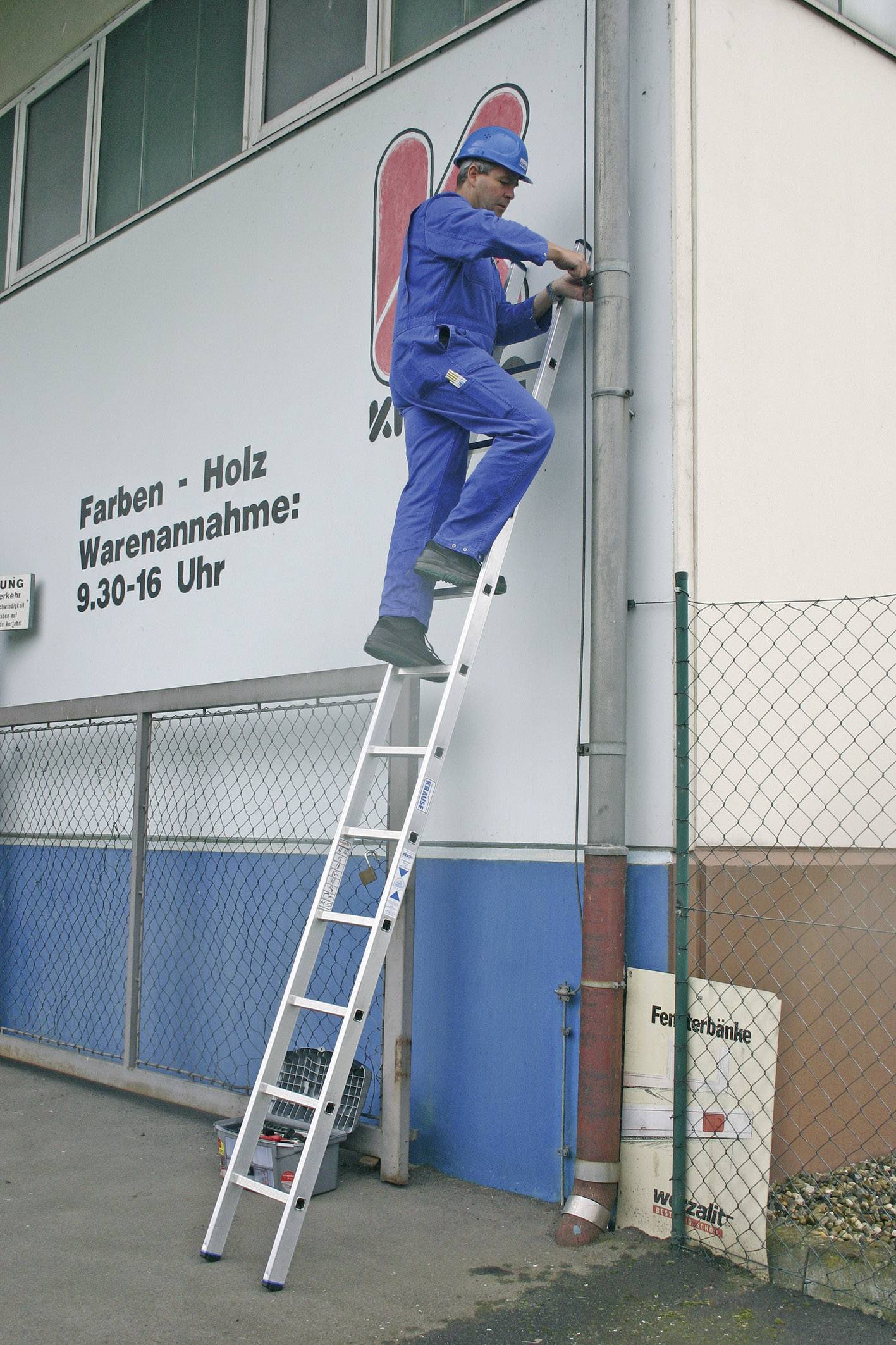 Eine Person in blauer Arbeitskleidung steht auf einer Leiter und arbeitet an einer Gebäudewand neben einem Schild mit Öffnungszeiten.
