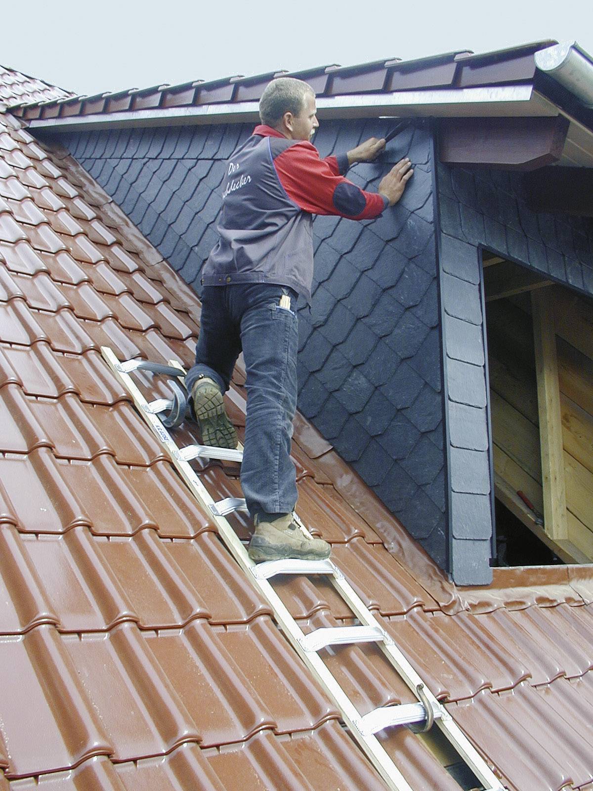 Ein Handwerker auf einer Dachleiter repariert Schieferplatten an einem Giebel. Dach mit roten Ziegeln und blauem Himmel im Hintergrund.