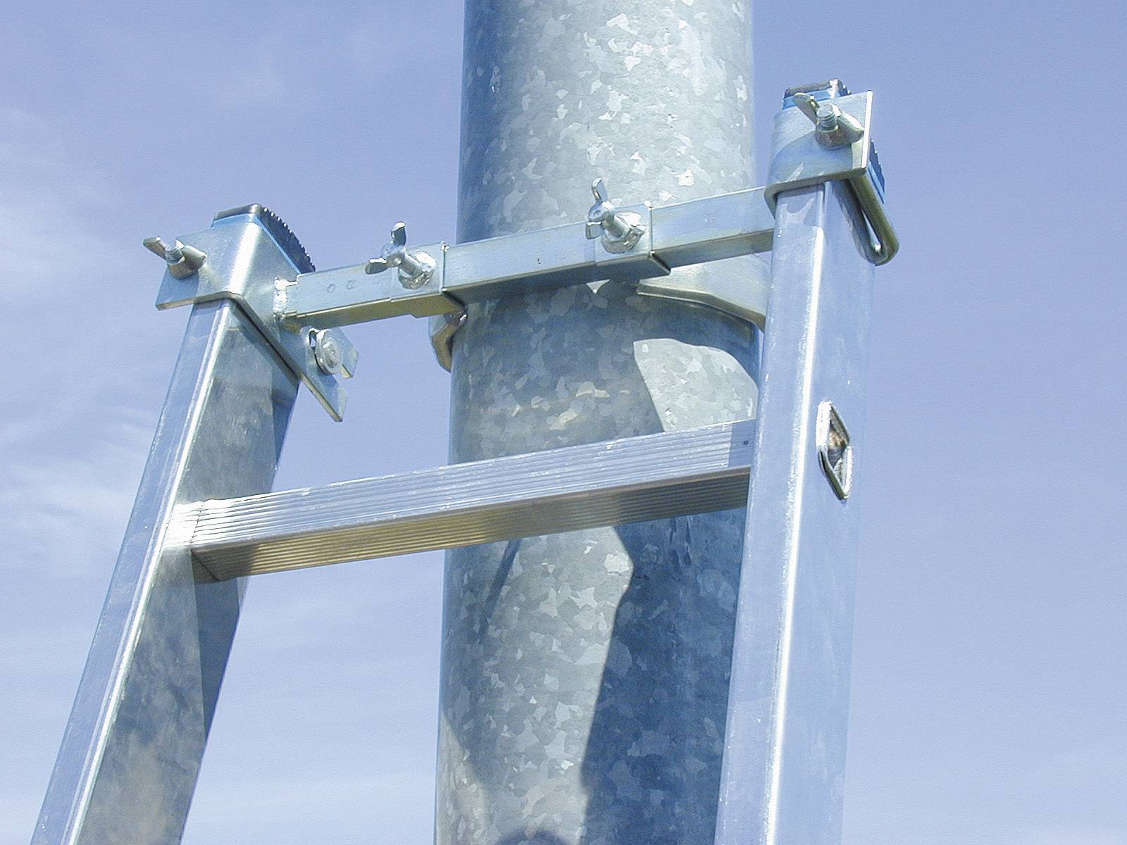 Eine silberne Metallleiter, die oben an einem runden, metallenen Pfosten mit Klammern befestigt ist, vor einem blauen Himmel.