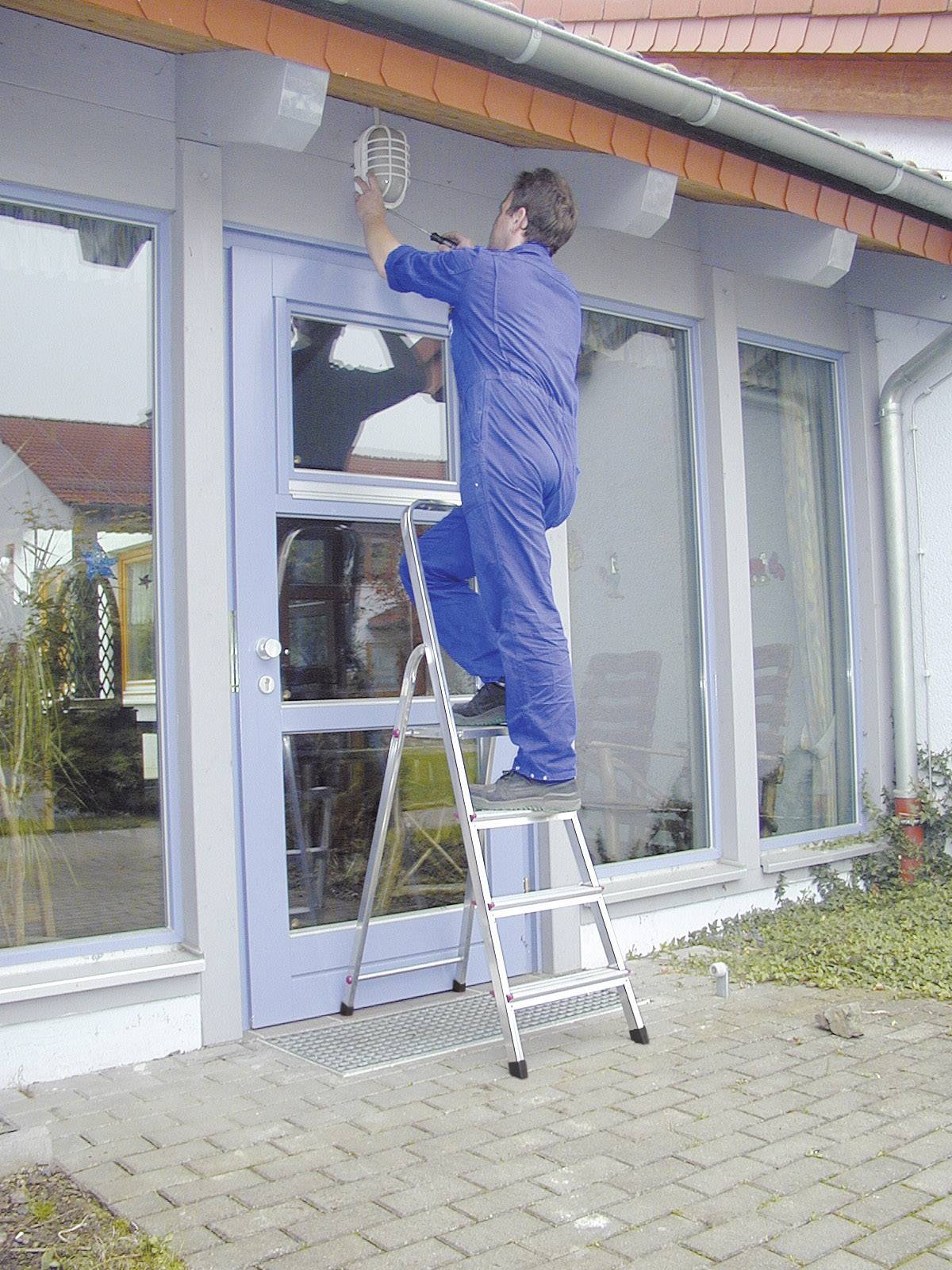 Ein Mann in blauer Arbeitskleidung steht auf einer Leiter und repariert eine Außenlampe über einer Eingangstür eines Gebäudes.