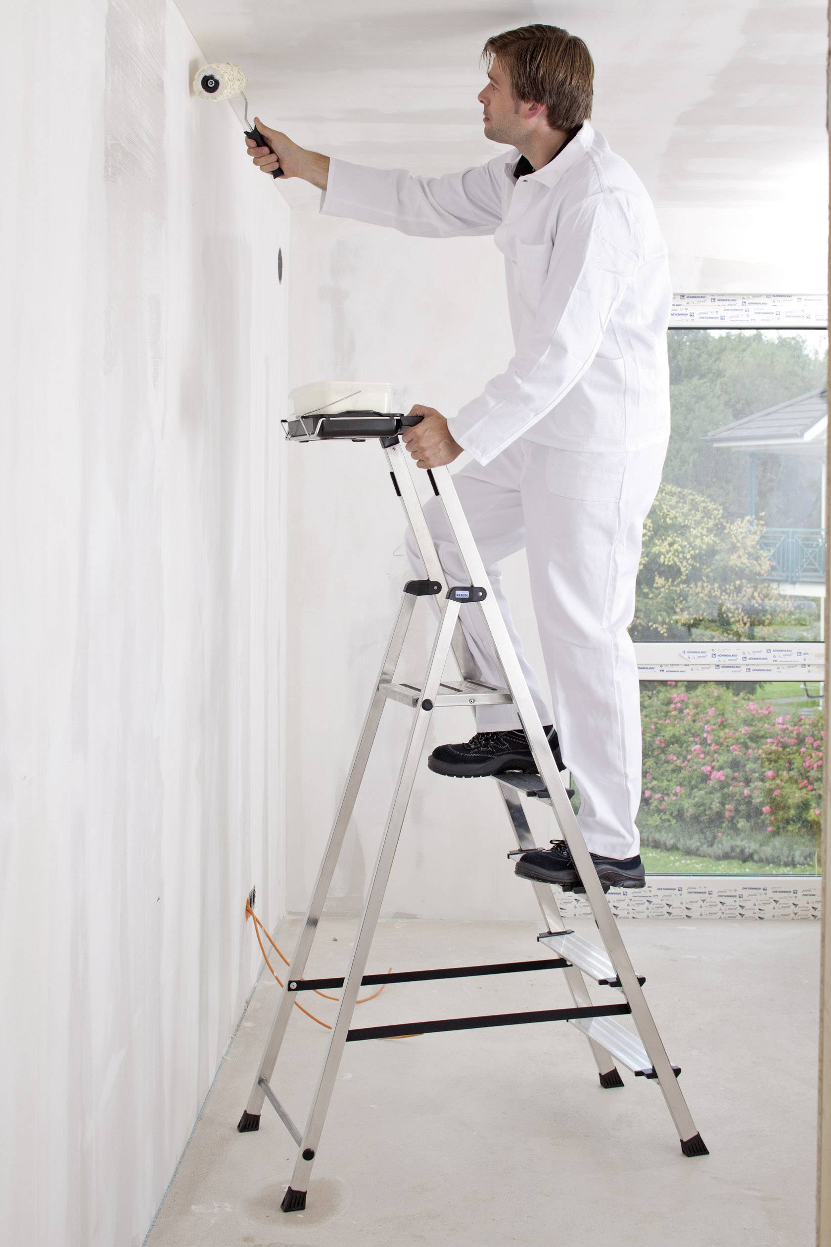 Ein Maler in weißer Arbeitskleidung steht auf einer Leiter und streicht eine weiße Wand mit einer Rolle. Im Hintergrund ist ein Fenster.