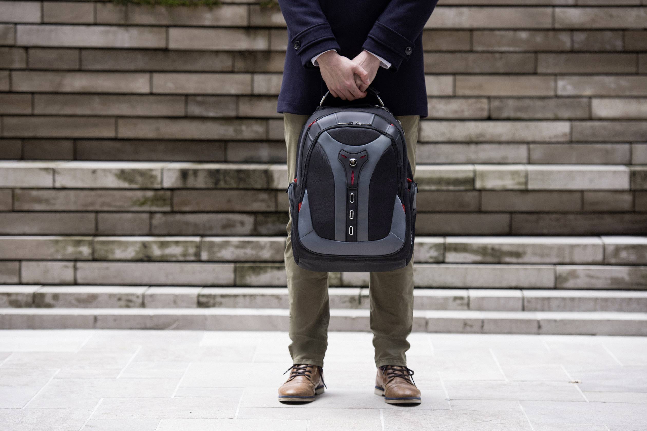 Eine Person hält einen Rucksack vor sich. Hintergrund mit Treppenstufen aus Stein. Die Person trägt braune Schuhe und einen dunklen Mantel.
